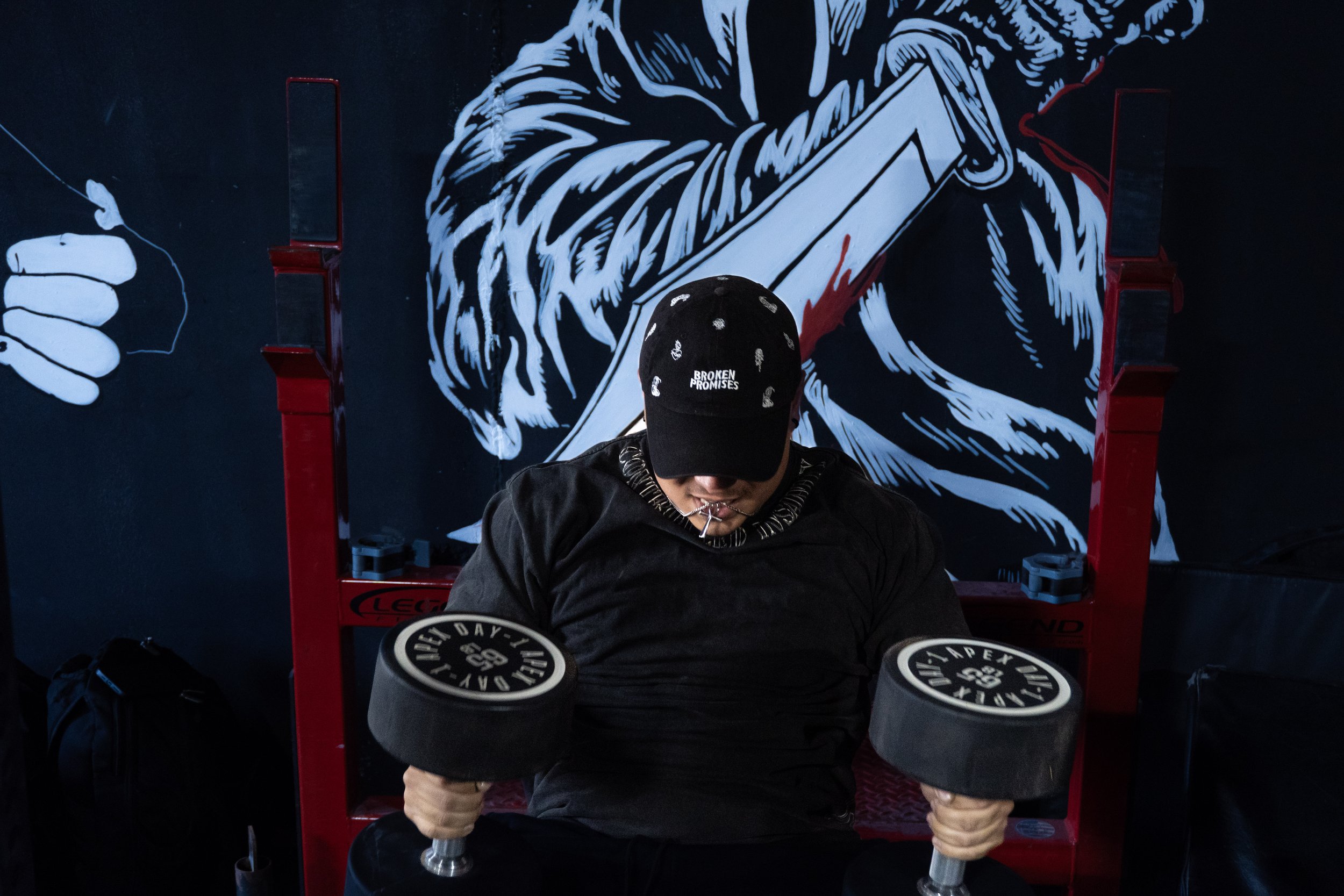A man seated with sunglasses and a black cap that says "Broken Promises" is lifting a dumbbell with black weights labeled "Day One" in a gym with a dark mural on the wall.