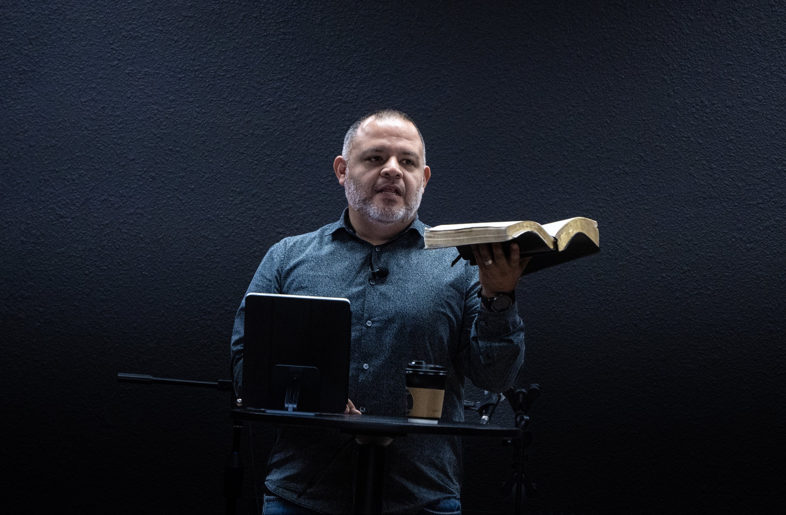 A man with a beard and short hair giving a presentation, holding an open Bible in one hand, standing in front of a dark textured wall with a table holding a laptop and a coffee cup.