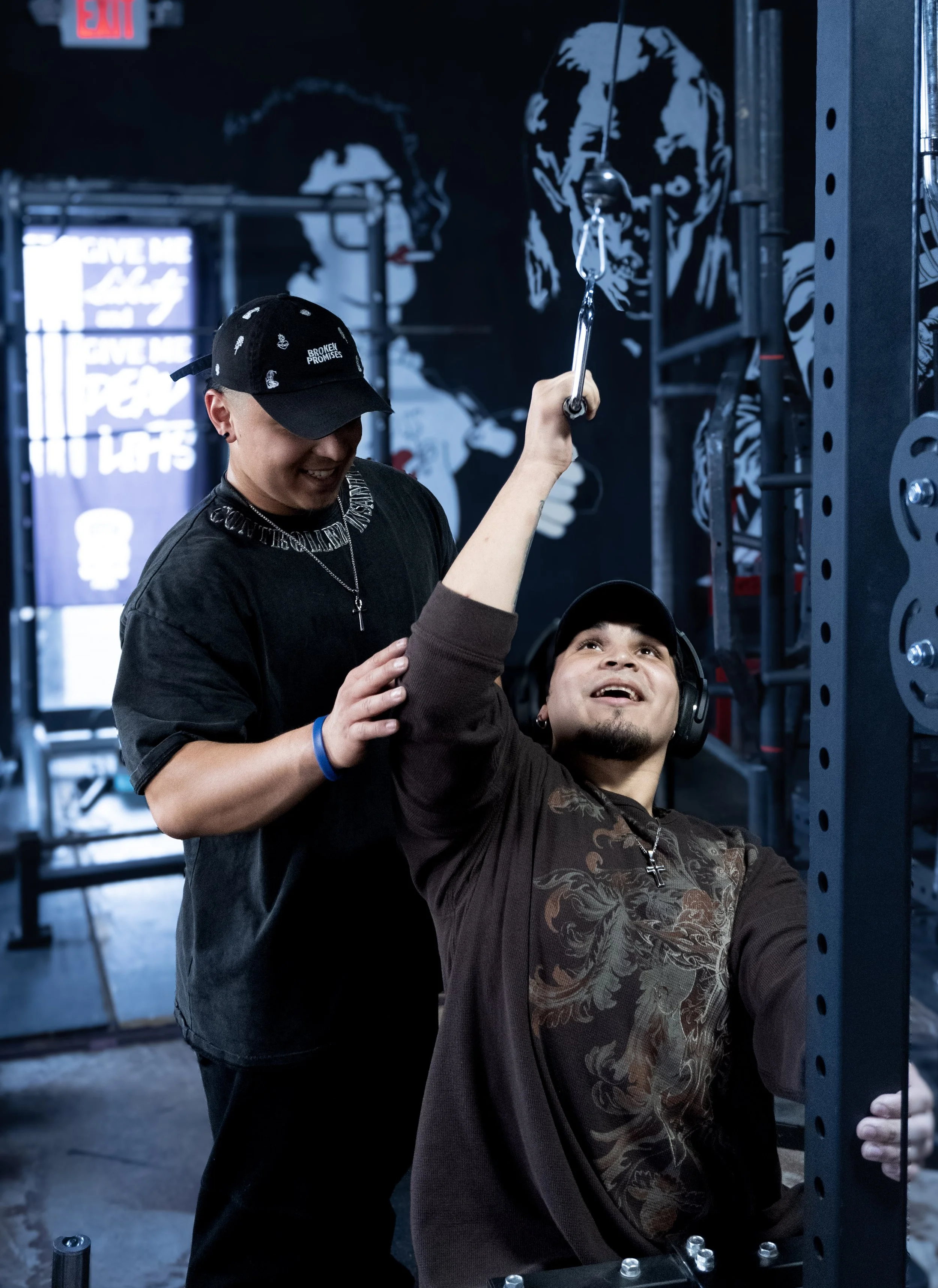 Two young men in a gym: one sitting on a bench pressing weights, wearing headphones and a cap, the other standing behind him, smiling and helping, in front of a mural of two women painted in black and white.