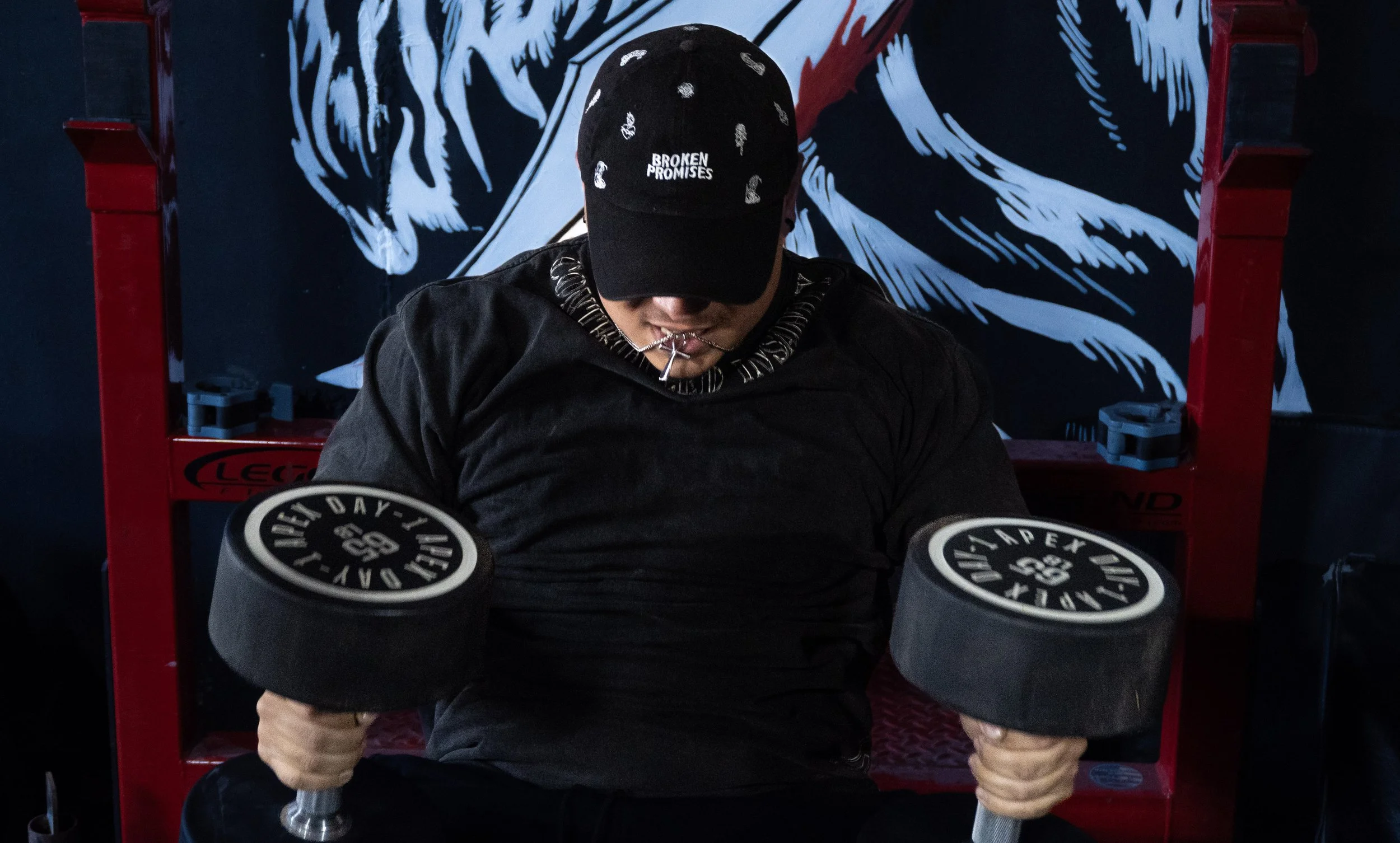 A man wearing a black hat with white text, a black hoodie, and holding two black weights, is sitting in front of a mural with blue and white abstract patterns.