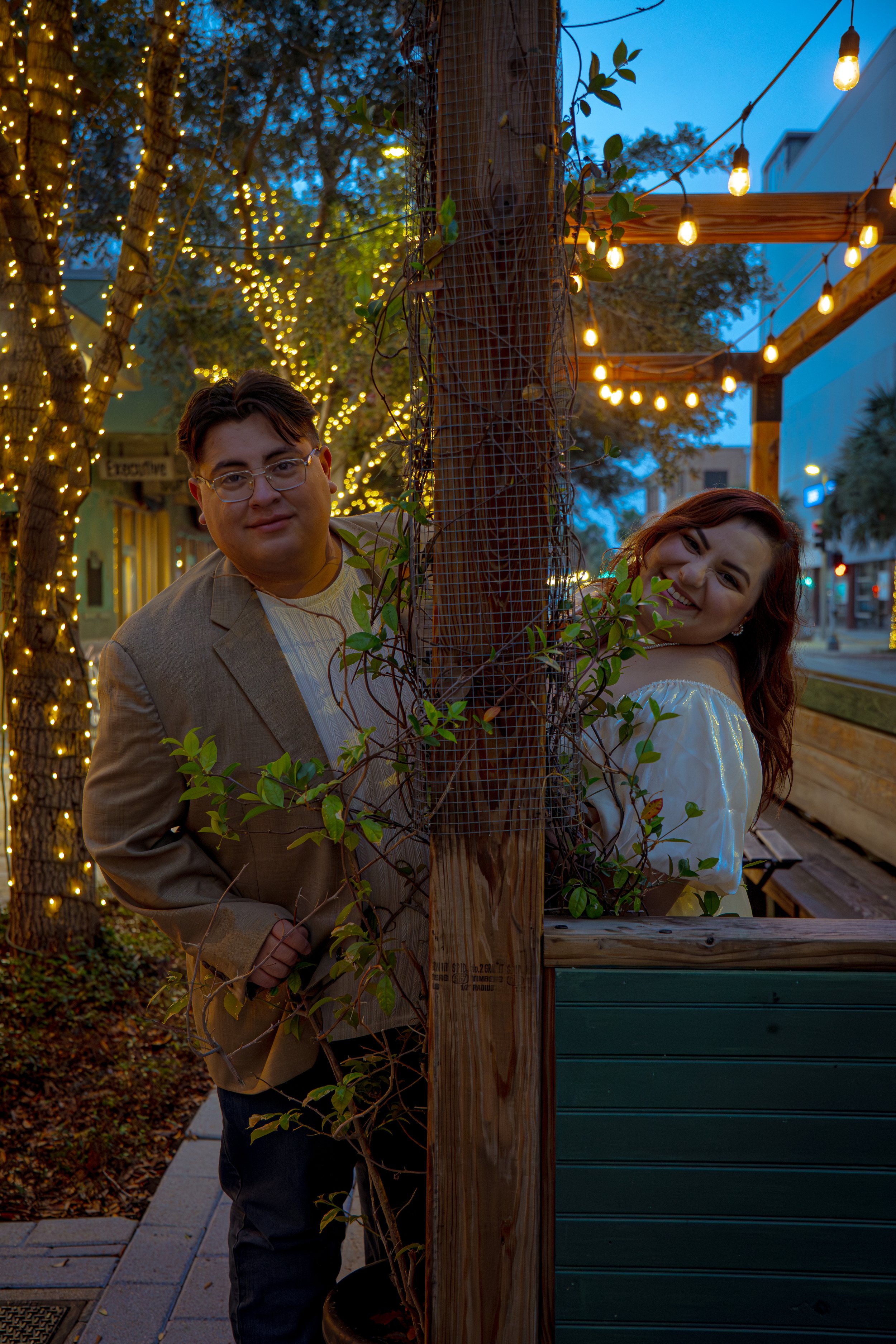 A man in glasses and a beige blazer standing next to a woman with red hair and a white dress, both leaning behind a wooden post with wire fencing and vines, in an outdoor setting with string lights and trees in the evening.