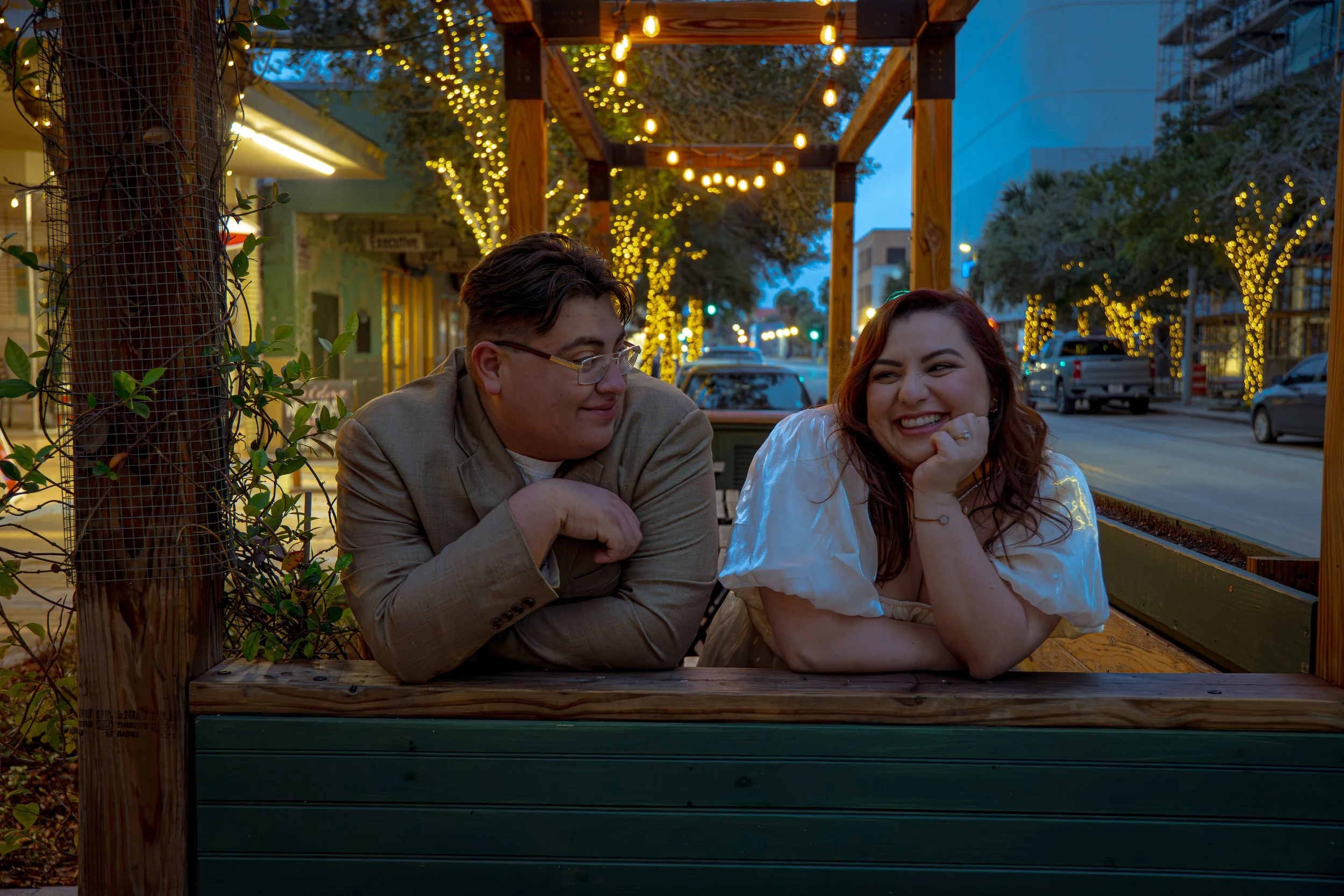 A young man and woman sit at an outdoor table at dusk, smiling and engaging in conversation. String lights are hanging overhead and trees are decorated with small lights, creating a warm, festive atmosphere in an urban setting.