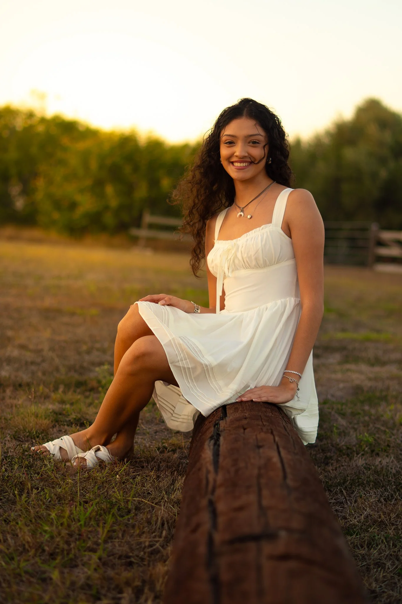 A young woman sitting on a log outdoors in a white dress at sunset, smiling at the camera.