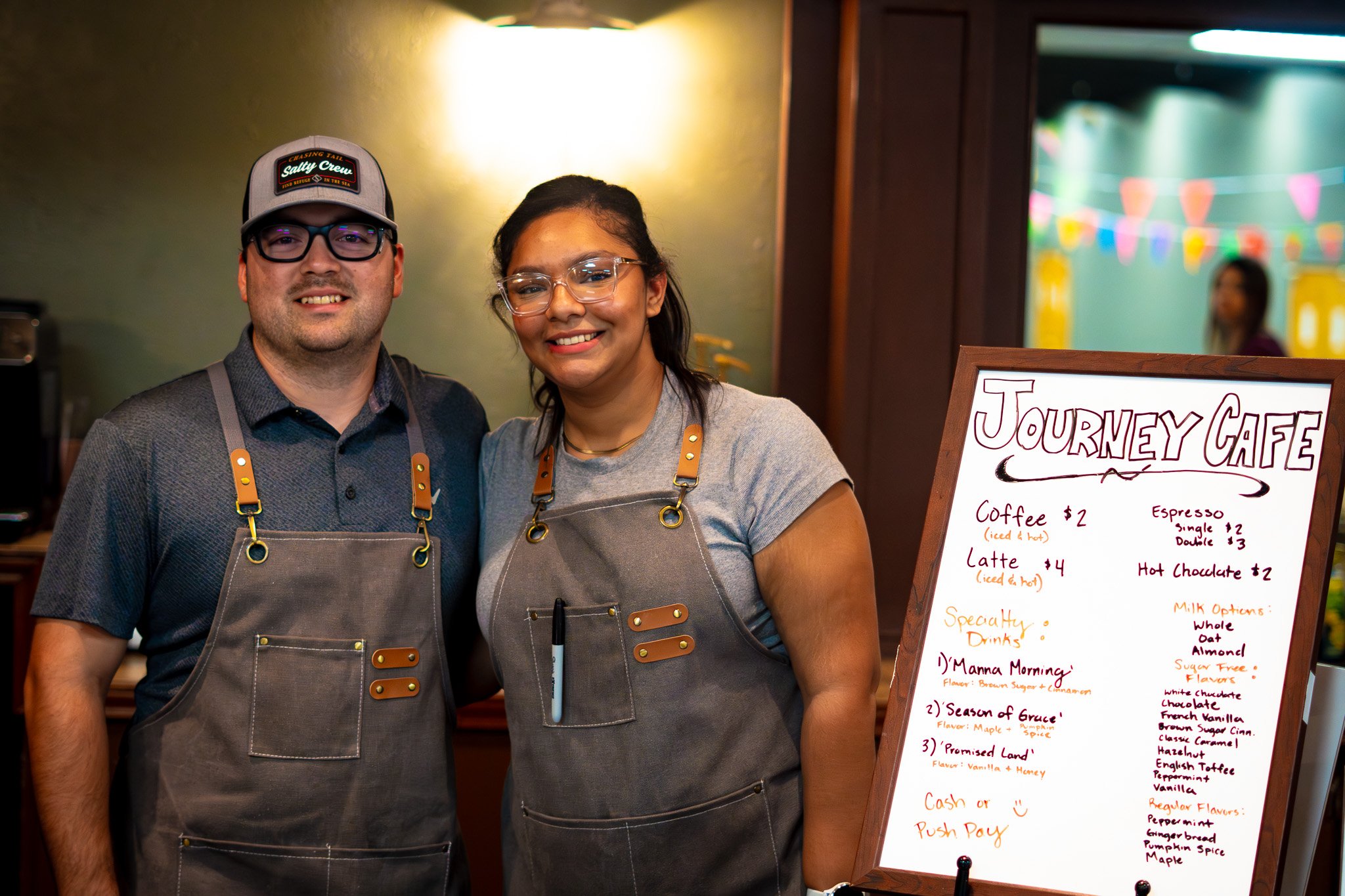 Two people standing inside a cafe, smiling at the camera, wearing aprons. There is a menu board beside them listing coffee and specialty drinks at Journey Cafe.