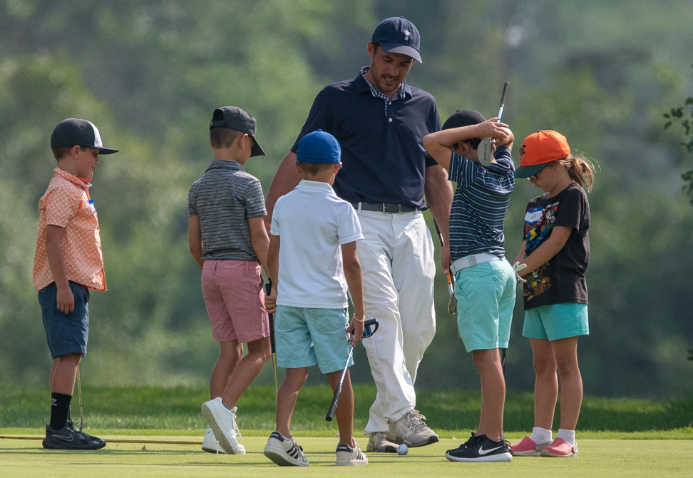 A golf instructor teaches five children how to putt on a golf course. The children are wearing colorful casual clothes and golf hats, with one girl in a black t-shirt and teal shorts, and a boy in an orange cap.