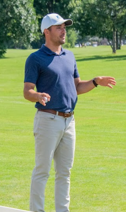 A man standing on a golf course, wearing a blue polo shirt, light-colored pants, and a white cap, gesturing with his hands.