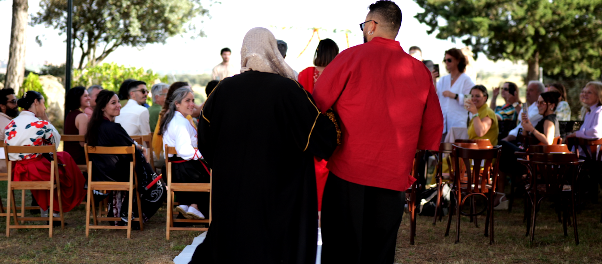 Una ceremonia al aire libre con varias personas sentadas en filas y otras de pie, varias tomadas con smartphones, bajo árboles verdes en un día soleado.