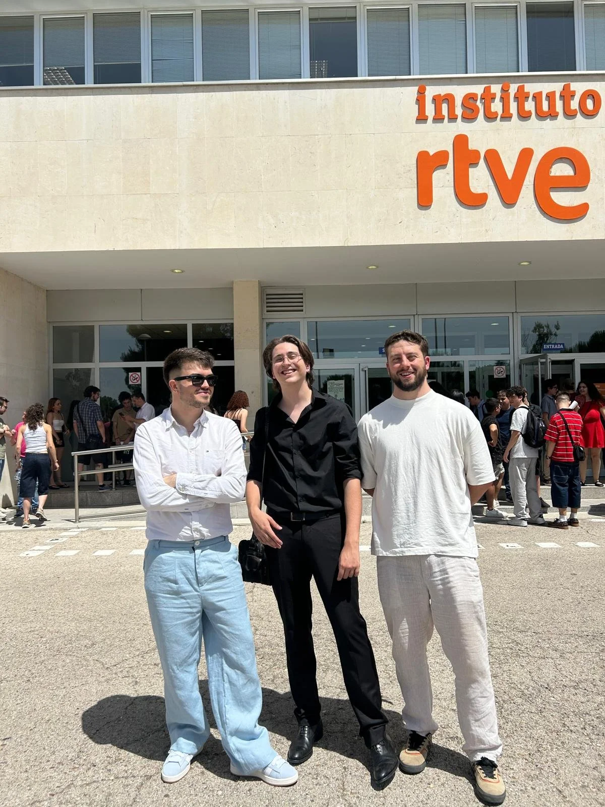 Tres personas de pie frente a un edificio con el logo 'instituto rtve', rodeadas de varias otras personas en la entrada del edificio.