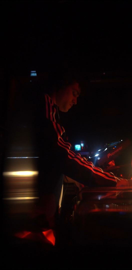 Persona mezclando música en un ambiente oscuro con equipo de DJ, luces rojas y negras.