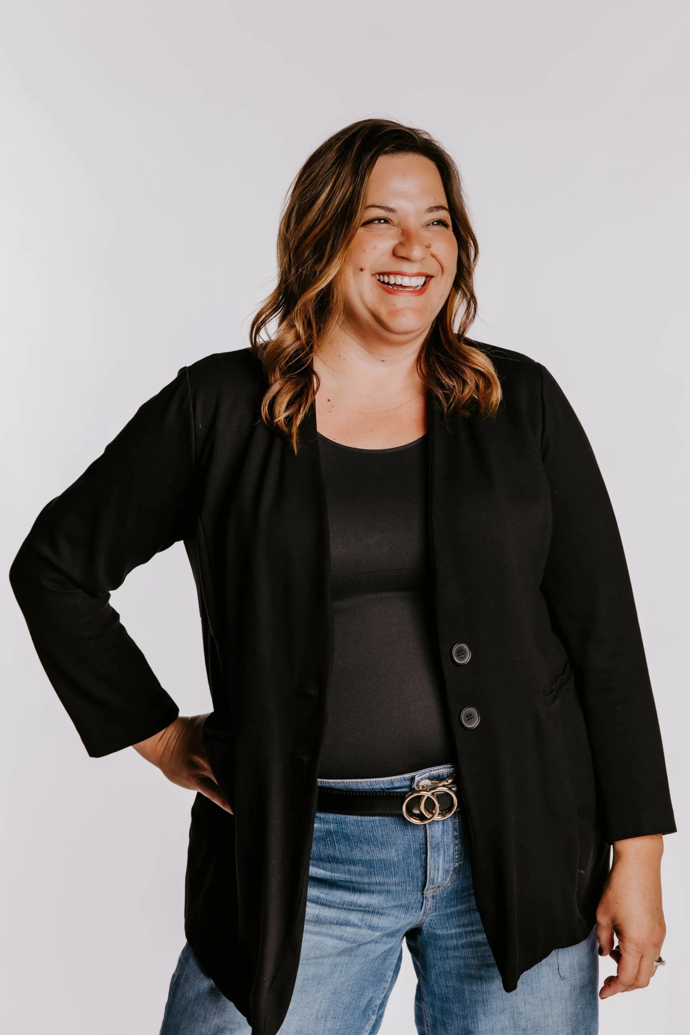 A woman with shoulder-length wavy brown hair, wearing a black blazer over a black top, blue jeans, and a black belt with silver rings, smiling and standing against a plain white background.