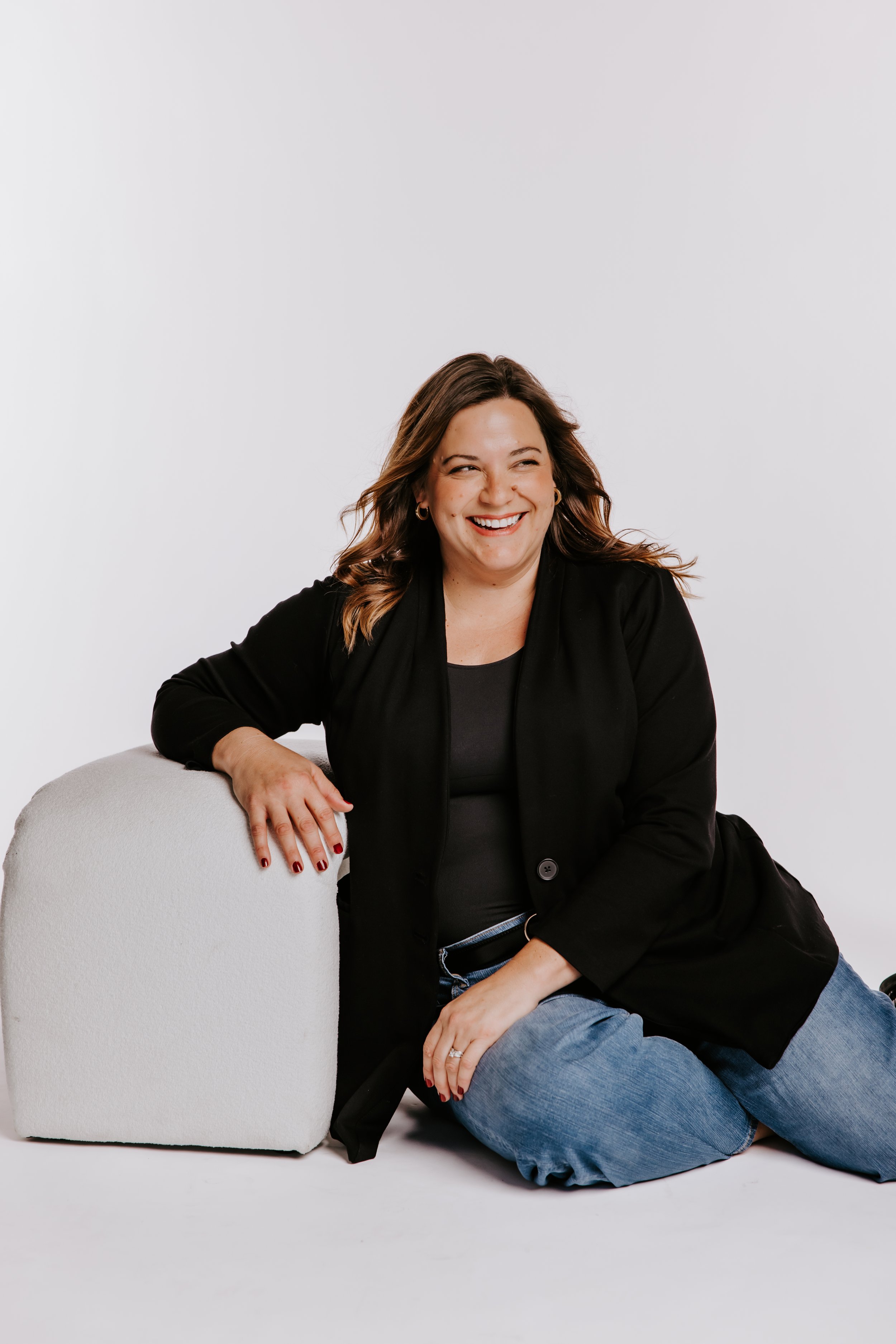 A woman with shoulder-length brown hair, wearing a black blazer and jeans, sitting on the floor and leaning on a white rounded cube, smiling and looking to her right against a plain white background.