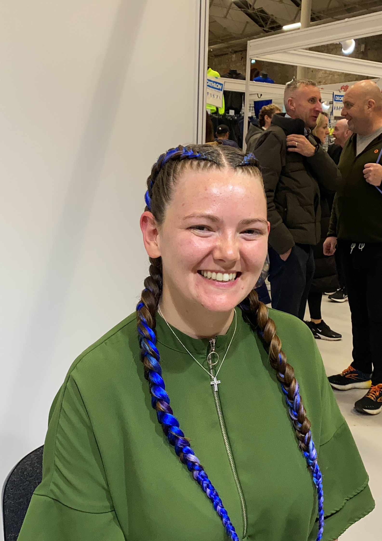 A young woman with braids, some dyed blue, smiling at the camera, wearing a green jacket and a cross necklace. In the background, people are engaged in conversation at an indoor event.