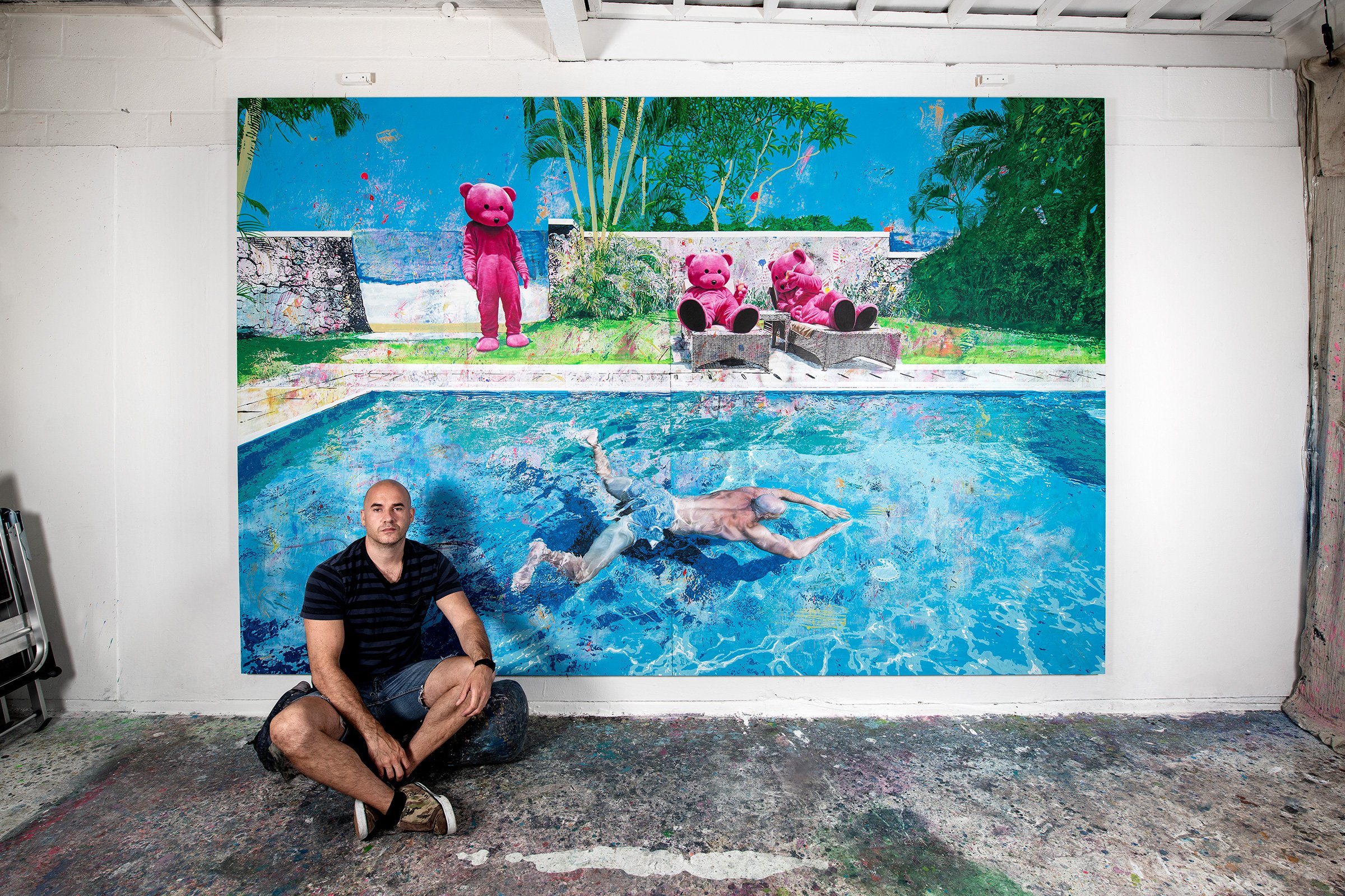 Artist LUAP - Paul Robinson - Artist sitting on the floor in front of a colorful painting of a person swimming in a pool with three pink teddy bears in the background, on a white wall in an art studio.