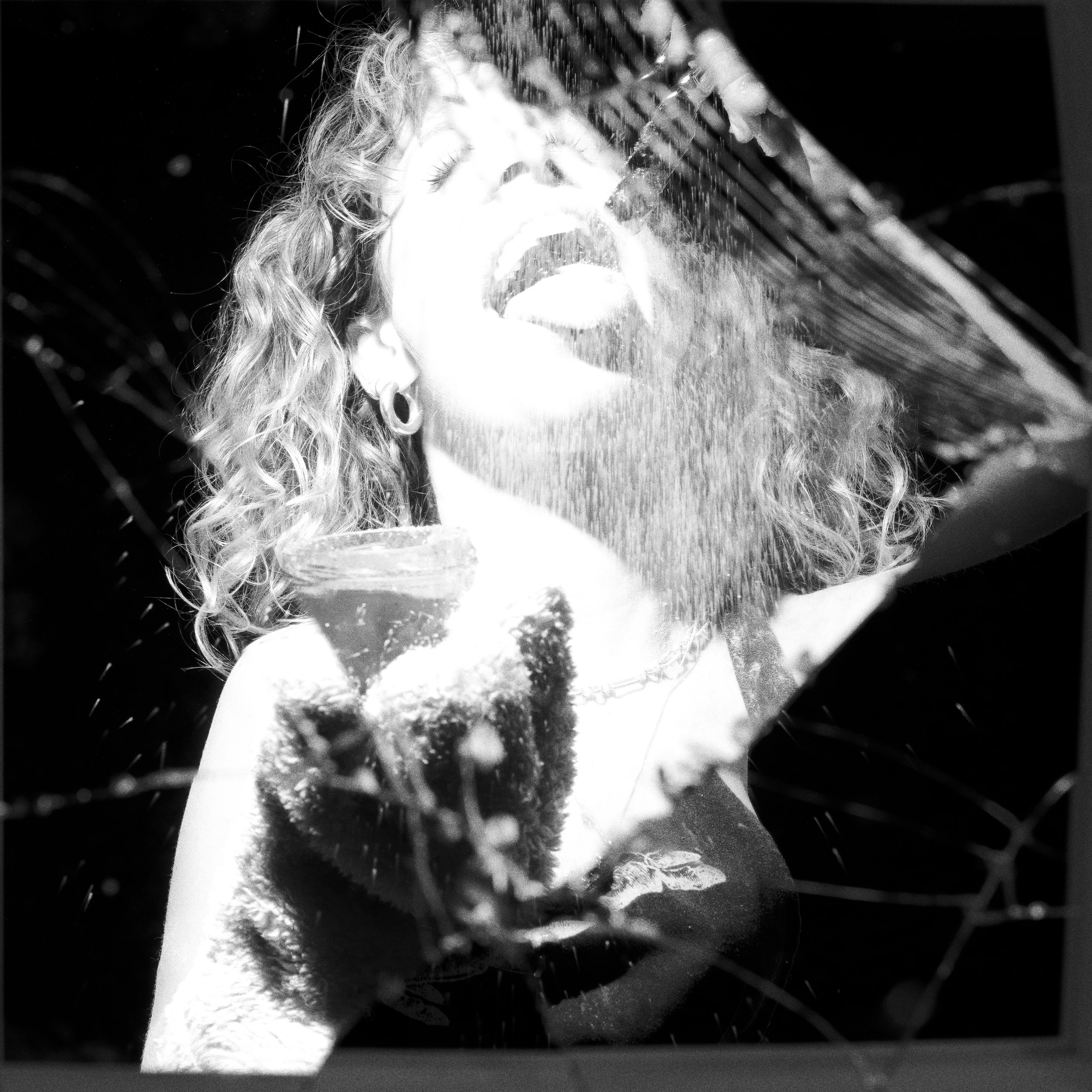 A woman with curly hair and earrings is smiling with her mouth open, holding a glass, as water splashes around her in black and white.