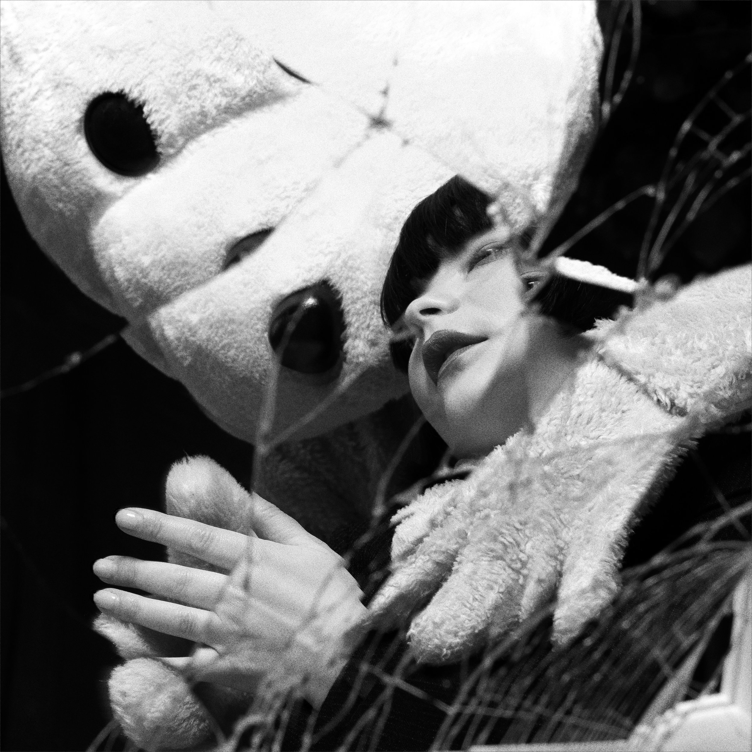 A woman with dark hair and lipstick lies close to a large plush teddy bear, surrounded by dried grass or leaves, creating a cozy and intimate scene in black and white.