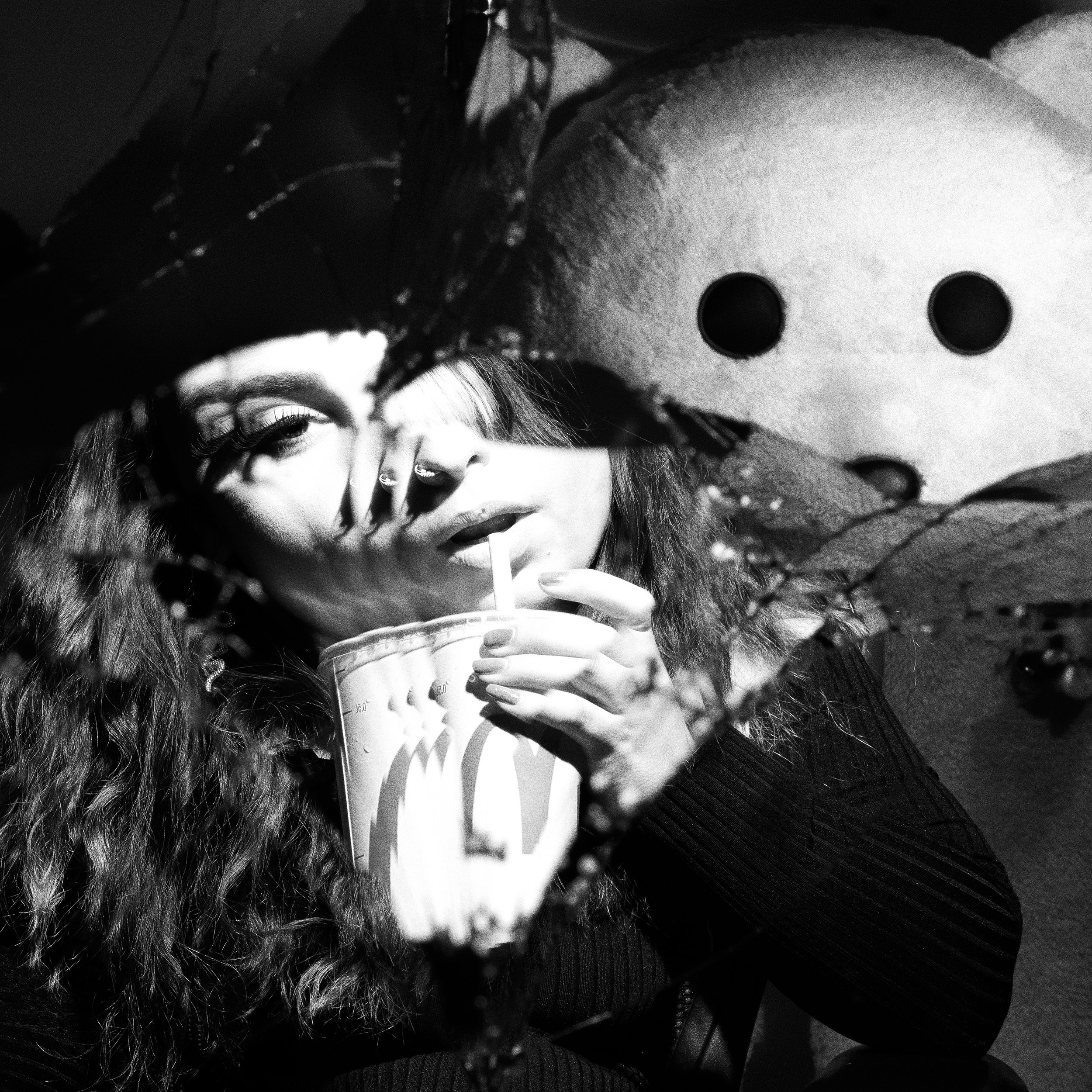 A woman with long wavy hair, holding a drink through a straw, poses in front of a broken mirror, with a teddy bear in the background.