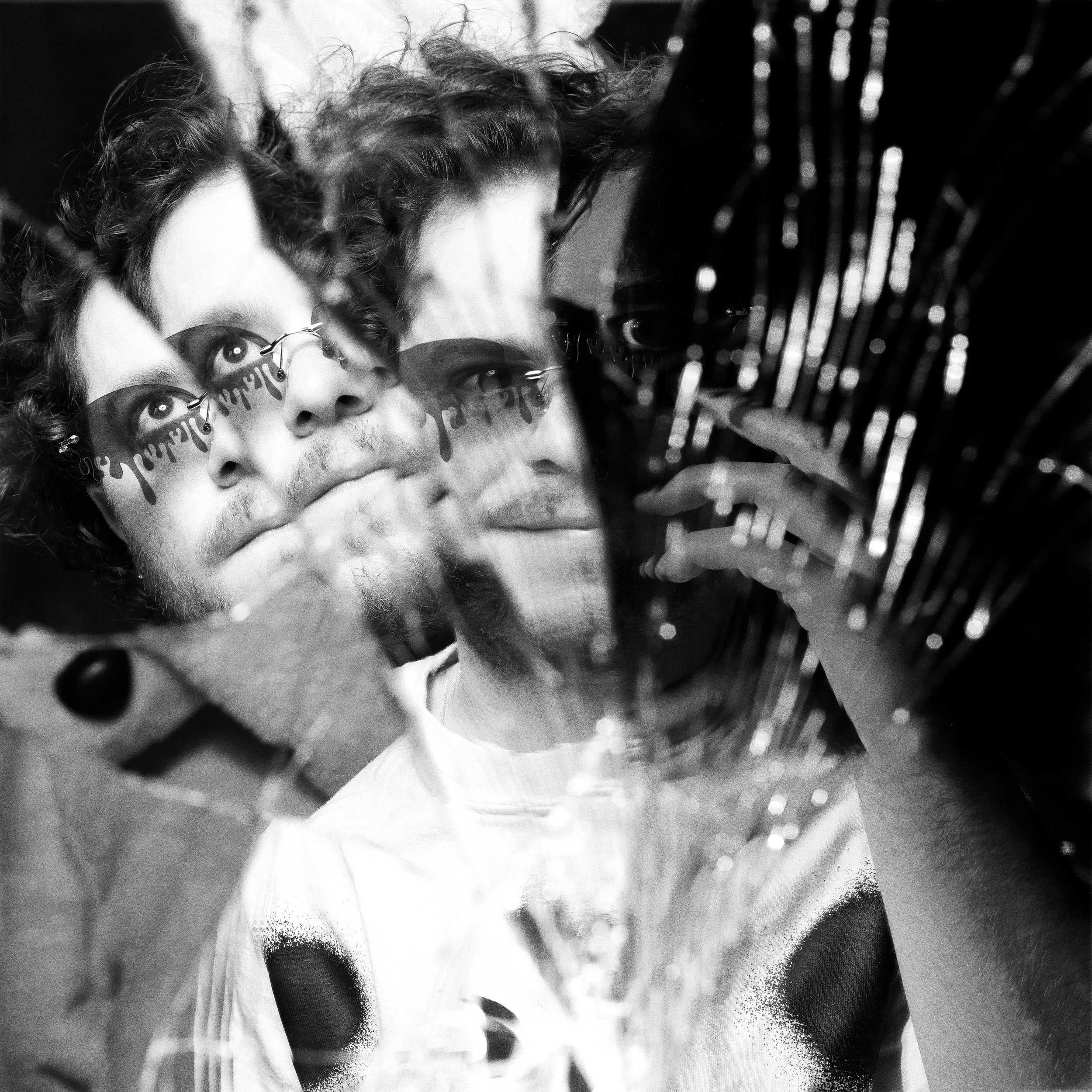 A person with curly hair and sunglasses looking into a reflective surface that creates multiple overlapping images of their face.