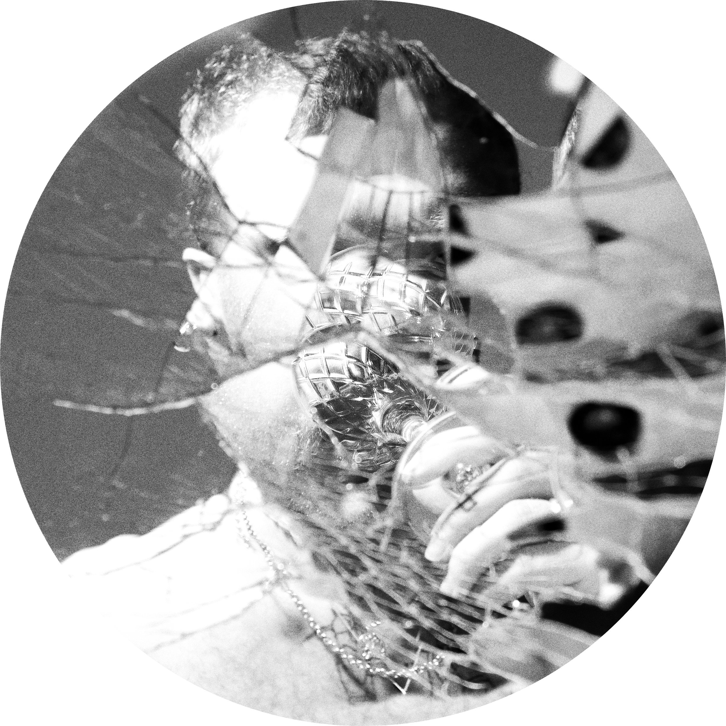 A black and white close-up photograph of a shiny, reflective object with multiple facets, possibly a piece of art or decorative item, with some human elements like a hand and part of a face visible in the reflection.