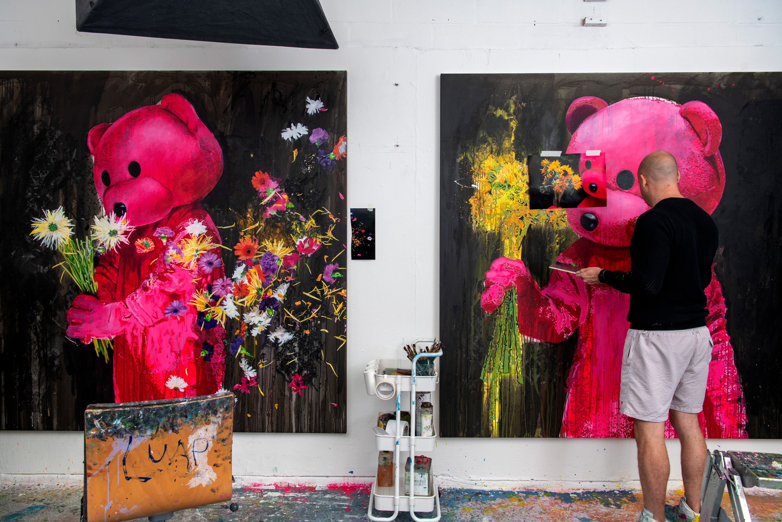 Artist LUAP - Paul Robinson - An artist stands in front of two large paintings of pink teddy bears on a gallery wall, with art supplies and paint splatters on the floor.