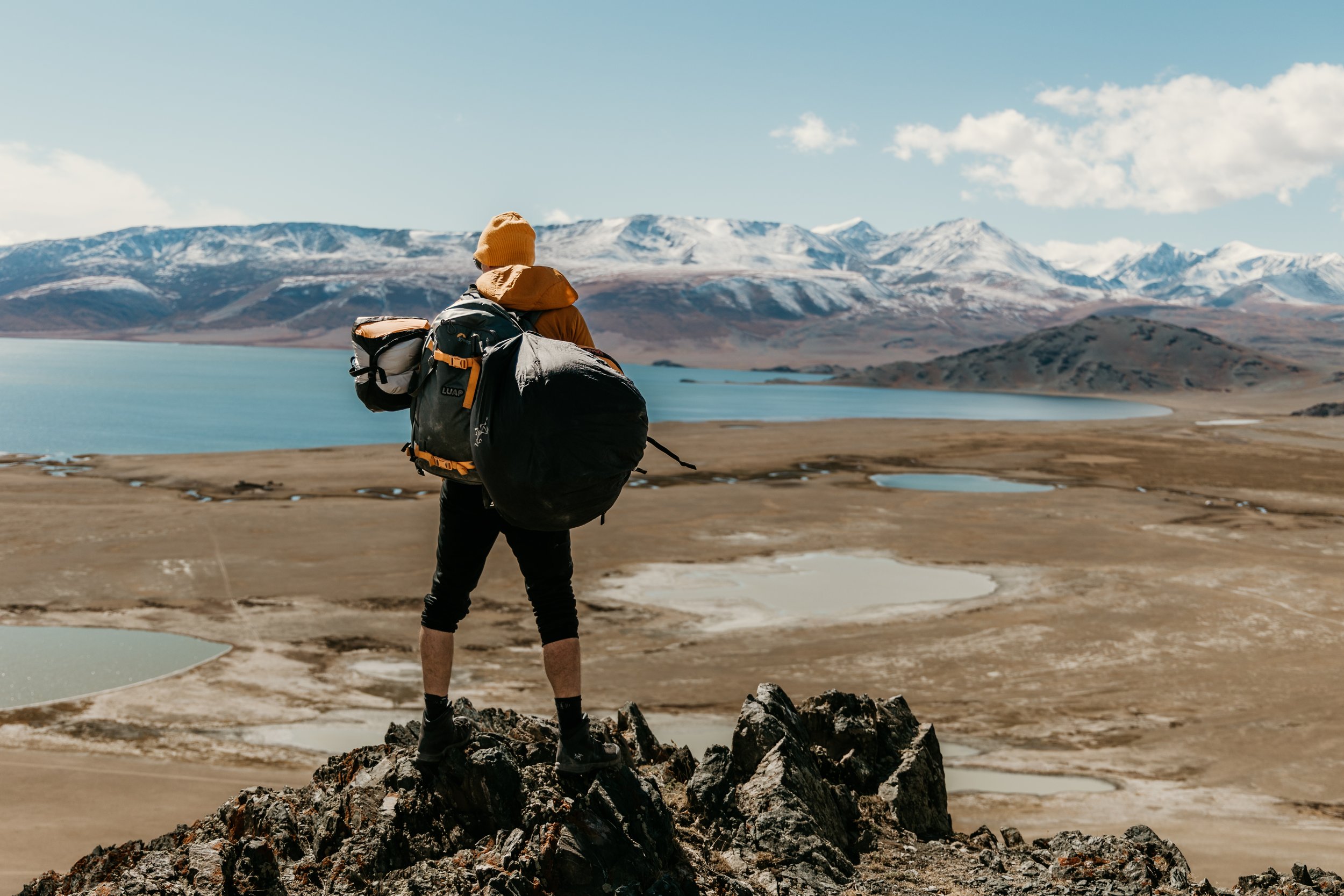Artist LUAP - Paul Robinson standing on rocks overlooking a mountain lake with snow-capped peaks in the distance.