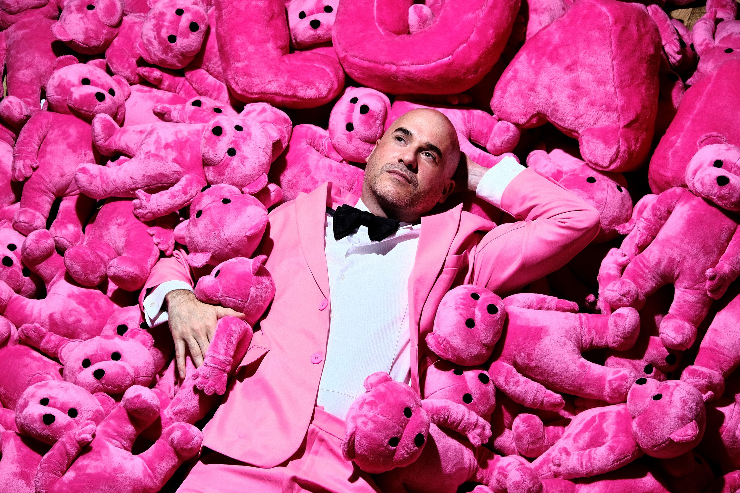 A man in a pink suit and black bow tie lying on a pile of pink teddy bears.