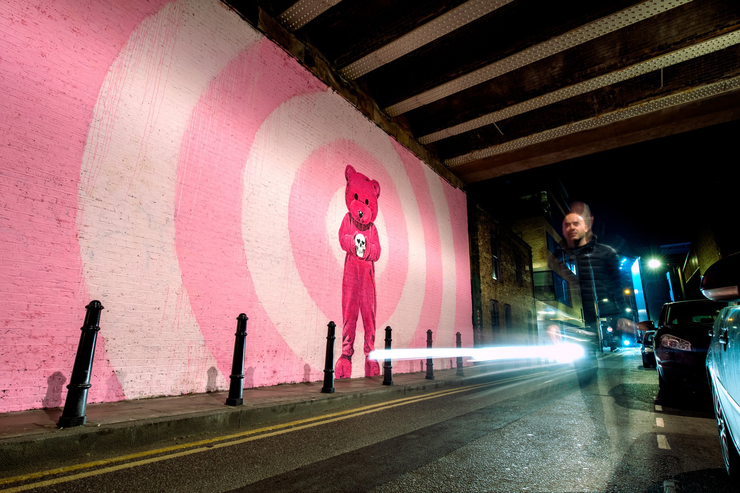 Artist LUAP - Paul Robinson - Street at night with a pink and white spiral mural of a teddy bear holding a skull, streetlights, cars, and a person passing by.