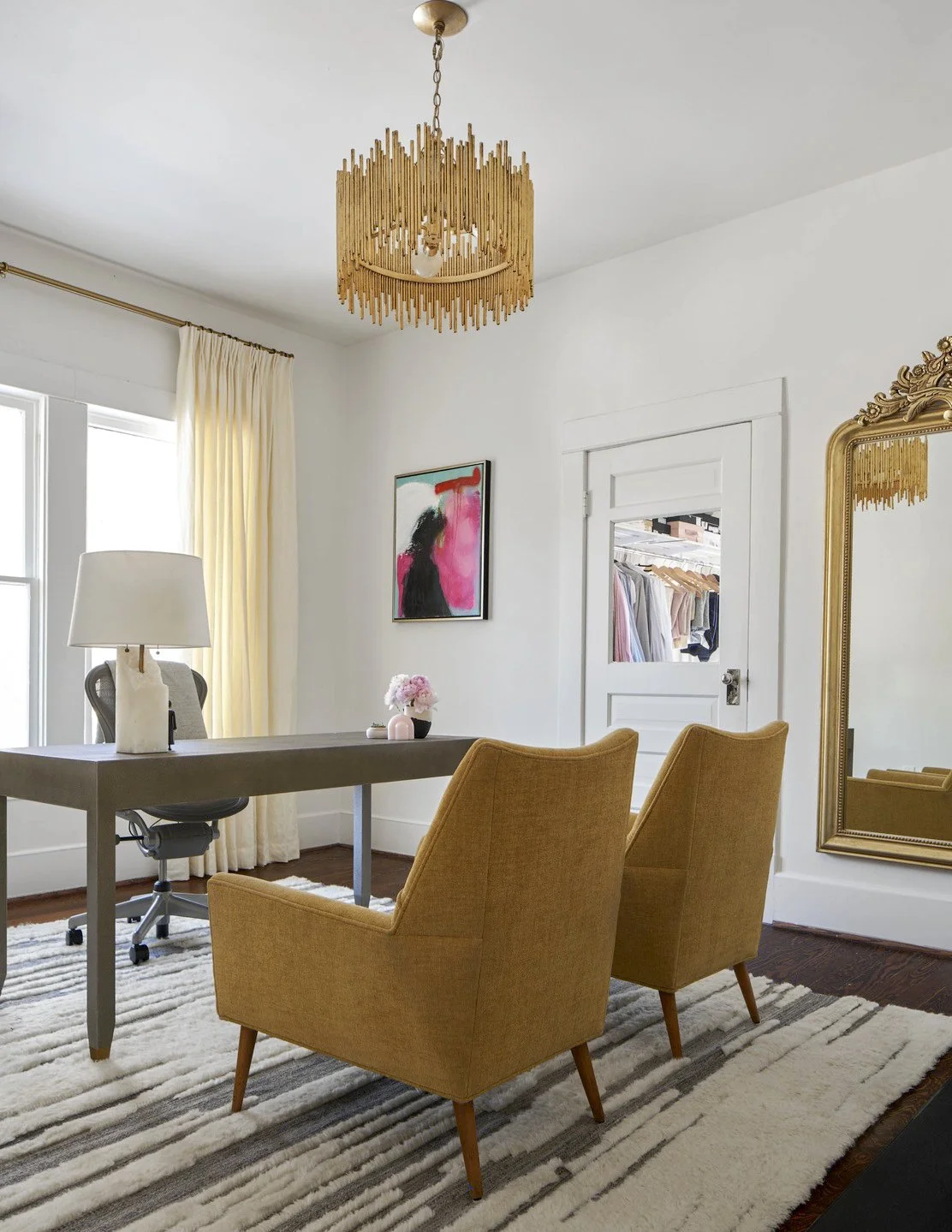 A home office with a gray desk, two mustard yellow armchairs, a large mirror with a gold frame, a white door with a small window showing a walk-in closet, a gold chandelier, a window with cream curtains, artwork featuring a pink, black, and white abstract silhouette, a table lamp with a cream base, and a vase with pink flowers on the desk.