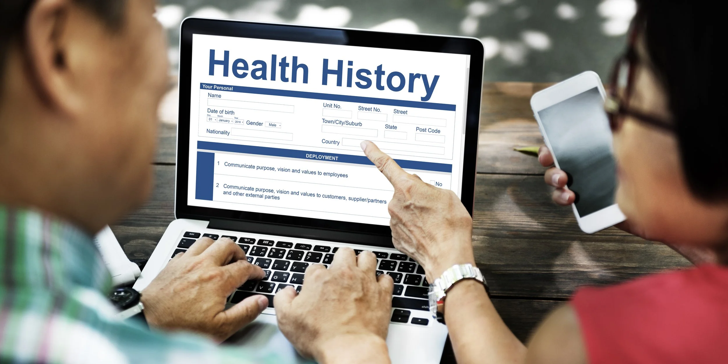 Two women reviewing a health history form on a laptop, with one pointing at the form and the other holding a smartphone.