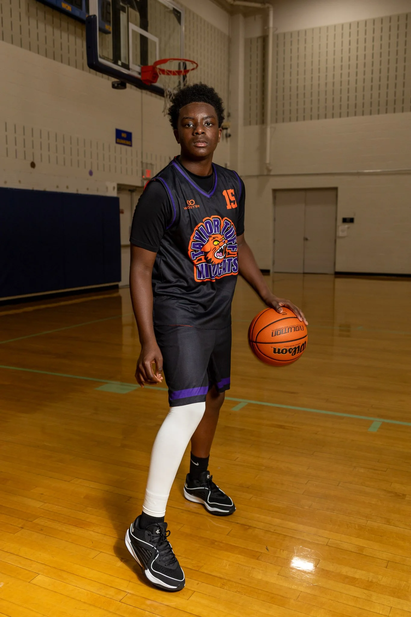 Young basketball player in a purple and orange team uniform holding a basketball and pointing at the camera in a gymnasium.
