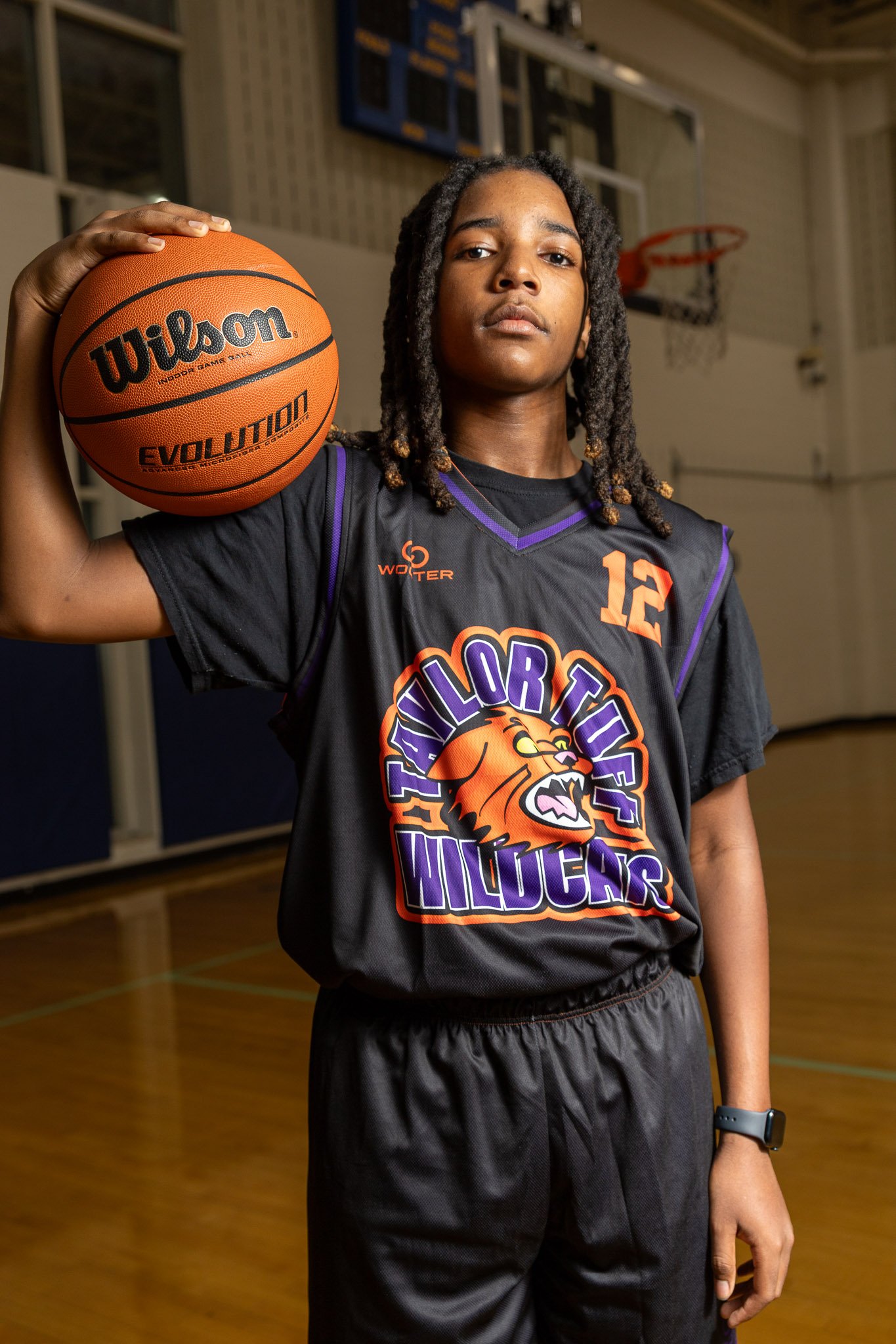 Basketball player holding two basketballs on his shoulders, standing inside a gym with a basketball hoop behind her. She is wearing a purple basketball jersey with a wildcat logo and the words "TAYLOR TUFF" and "WILDCATS."
