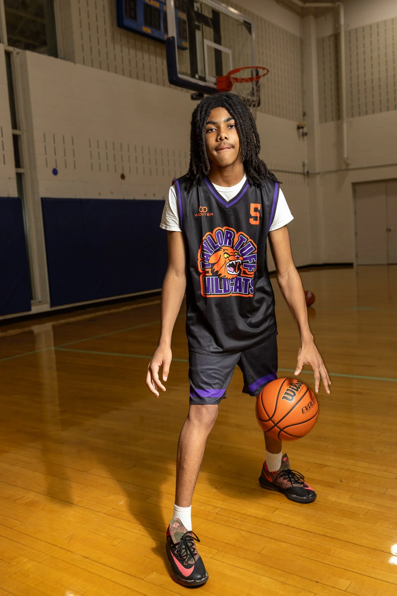 A young male basketball player with dreadlocks in a purple and orange team uniform, holding a basketball on an indoor court.
