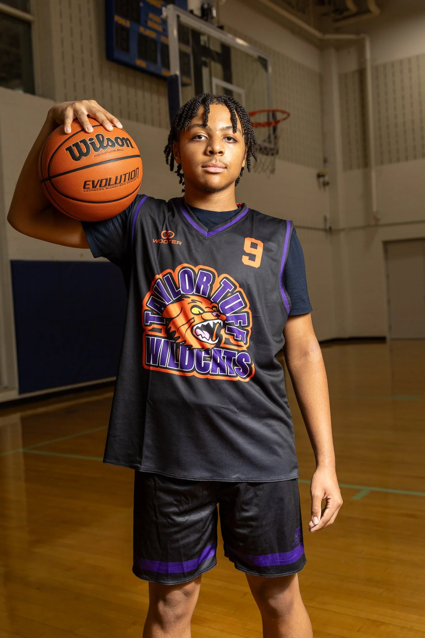 A young male basketball player in a purple jersey holding a Wilson basketball, standing in a gymnasium near a basketball hoop.