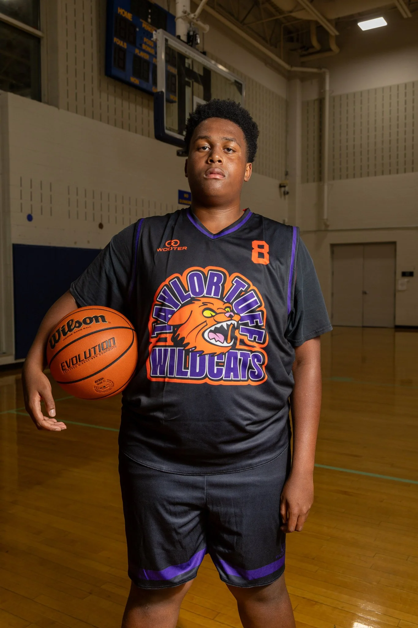 A young male basketball player wearing a purple and black team uniform holding a basketball on an indoor court.