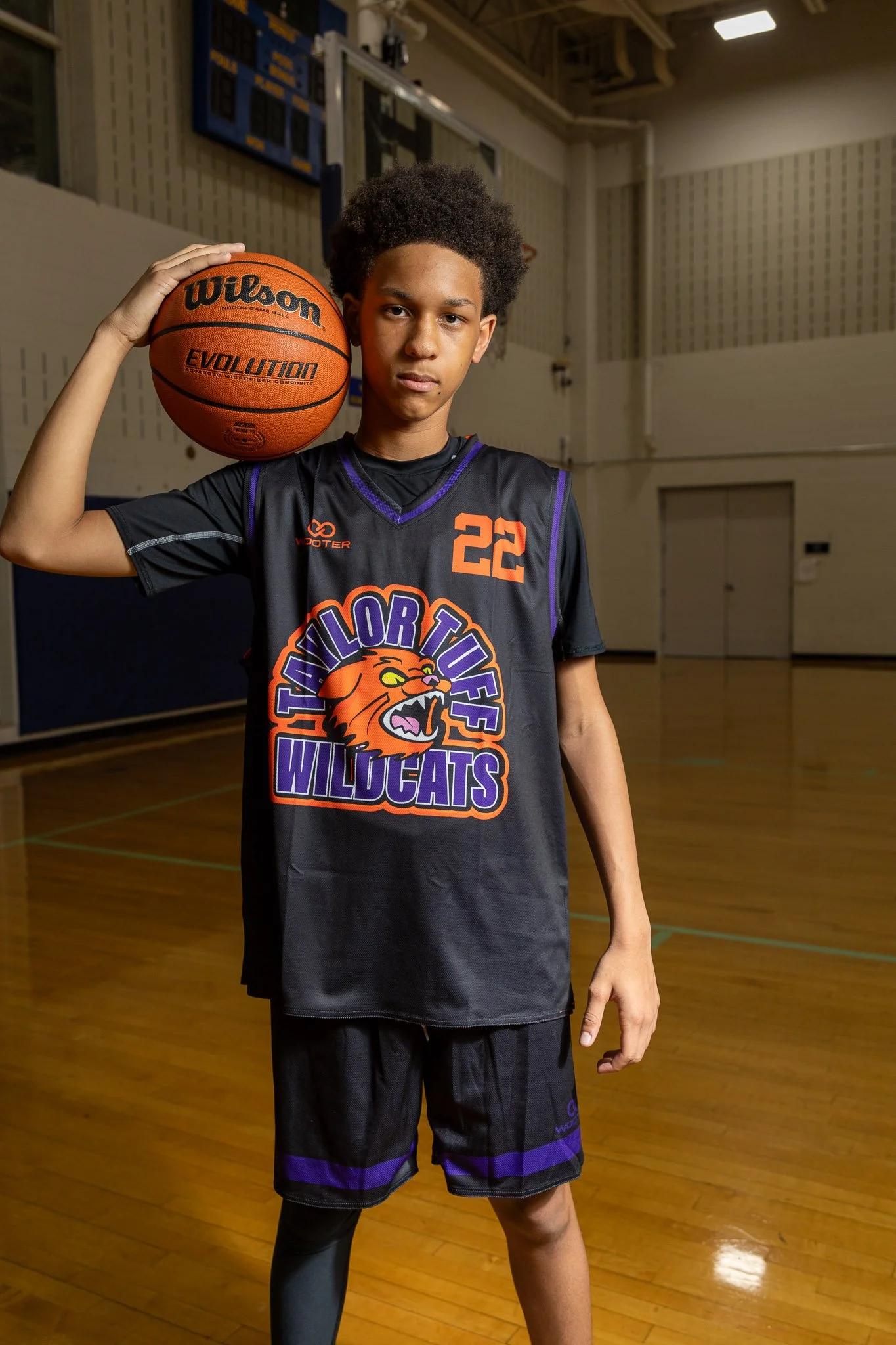A young male basketball player is standing on an indoor basketball court, holding a basketball and wearing a purple jersey with a wildcat logo, shorts, white sneakers, and black socks. The gym is lit with artificial lighting and has a basketball hoop and a 'No Dunking' sign in the background.