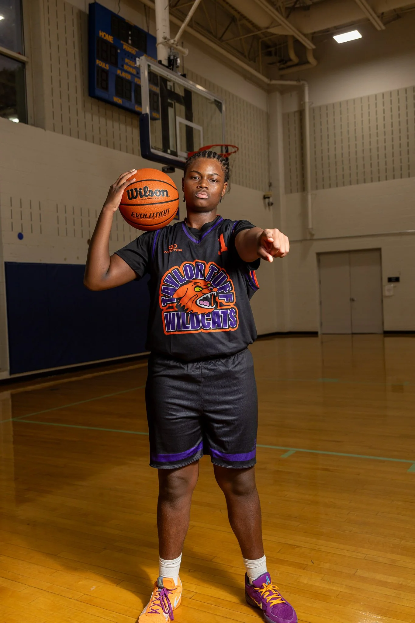 A young man with short curly hair standing indoors, wearing an orange athletic outfit and a black long-sleeve shirt with the logo 'Taylor Tuff Wildcats' featuring a wildcat's head.