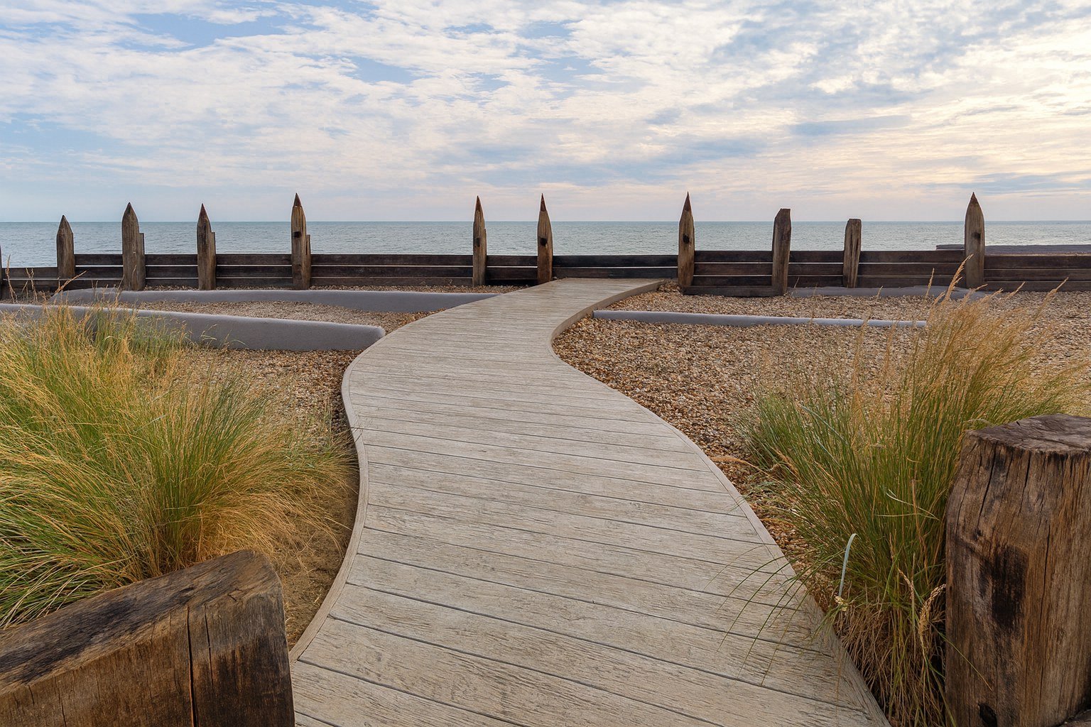A curved wooden pathway leads to a beach, bordered by plants and wooden posts, with a wooden fence overlooking the ocean under a cloudy sky.