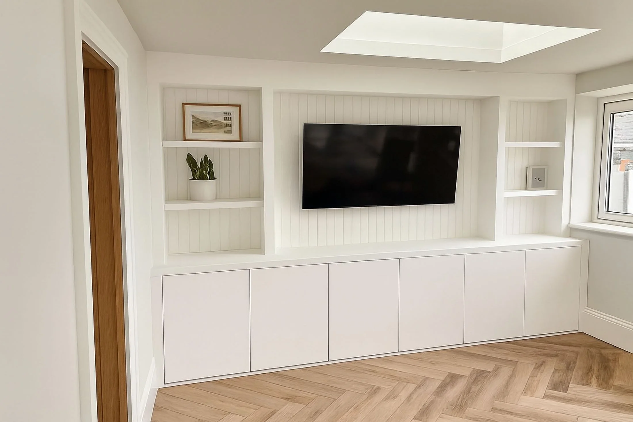 Modern living room with white built-in cabinetry, wall-mounted black flat-screen TV, a small framed picture on the wall, potted plant, window, and wooden flooring. Natural light from the skylight.