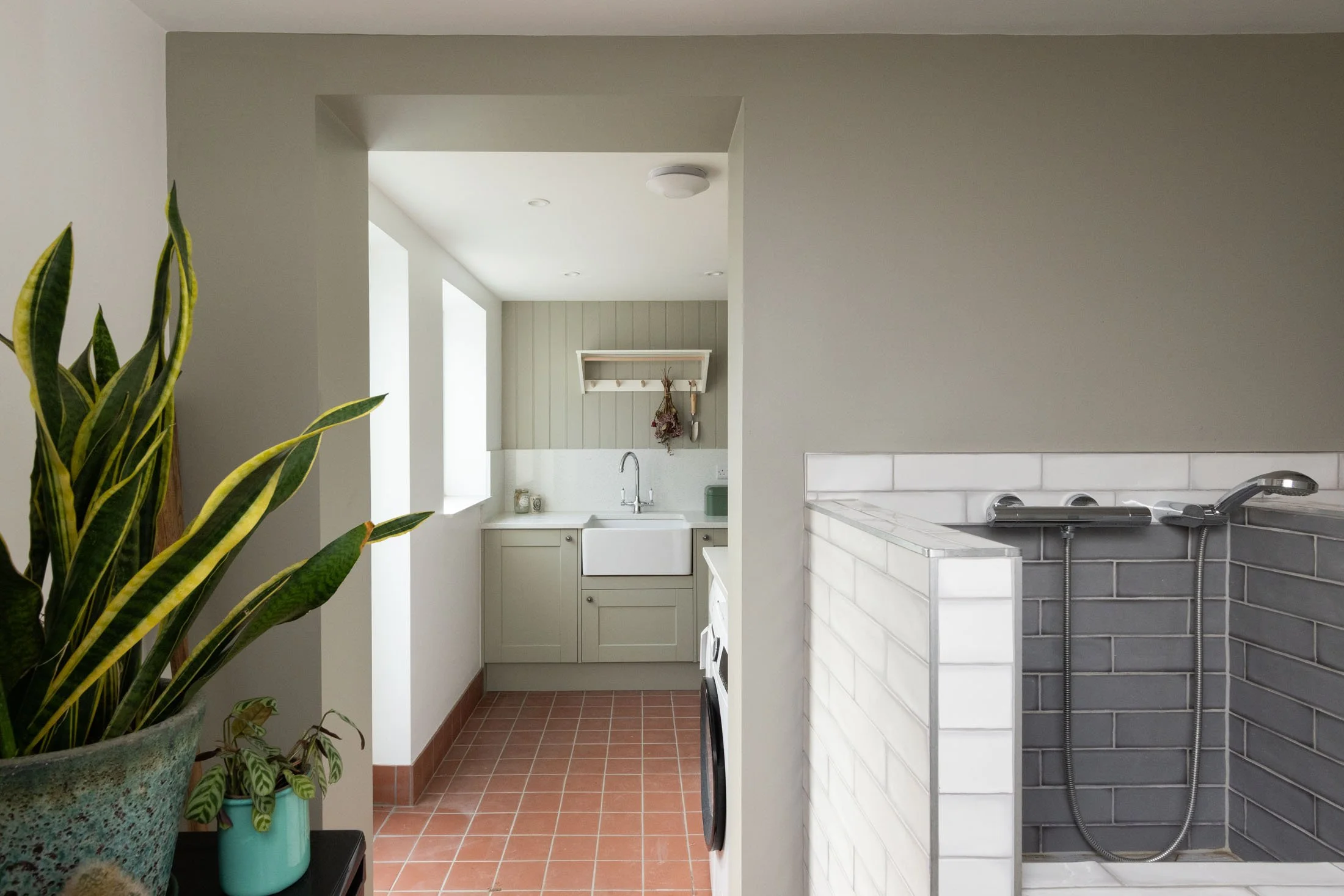 Interior view of a kitchen with white cabinets, a sink, potted plants on a black table, and a laundry area with a washing machine and dryer. The space has terracotta tile flooring and a neutral color palette.