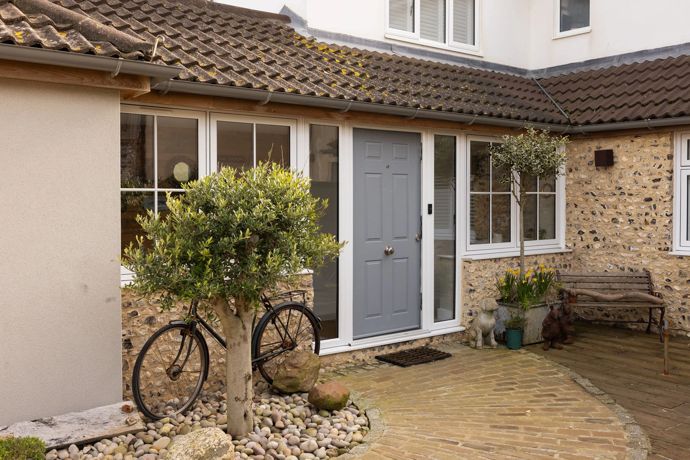 A house with a combination of stone and stucco exterior walls, a gray front door, and large glass windows. There are plants, including a small tree and yellow flowers, next to the house, along with a bike, rocks, and a cobblestone pathway leading to the door. The roof is tiled with some moss and lichen.