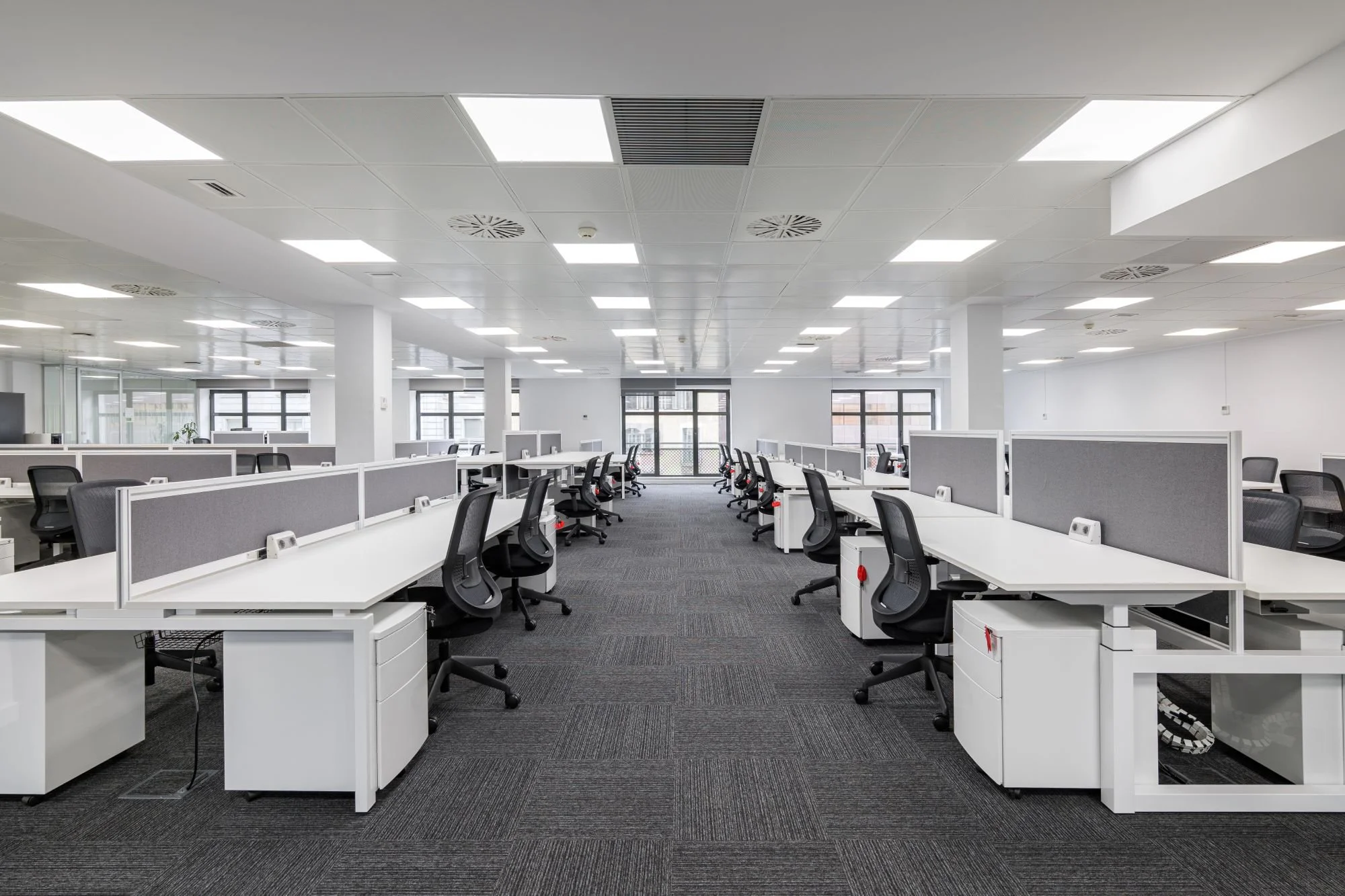 Empty modern office space with rows of white desks and black office chairs, illuminated by bright ceiling lights and large windows.
