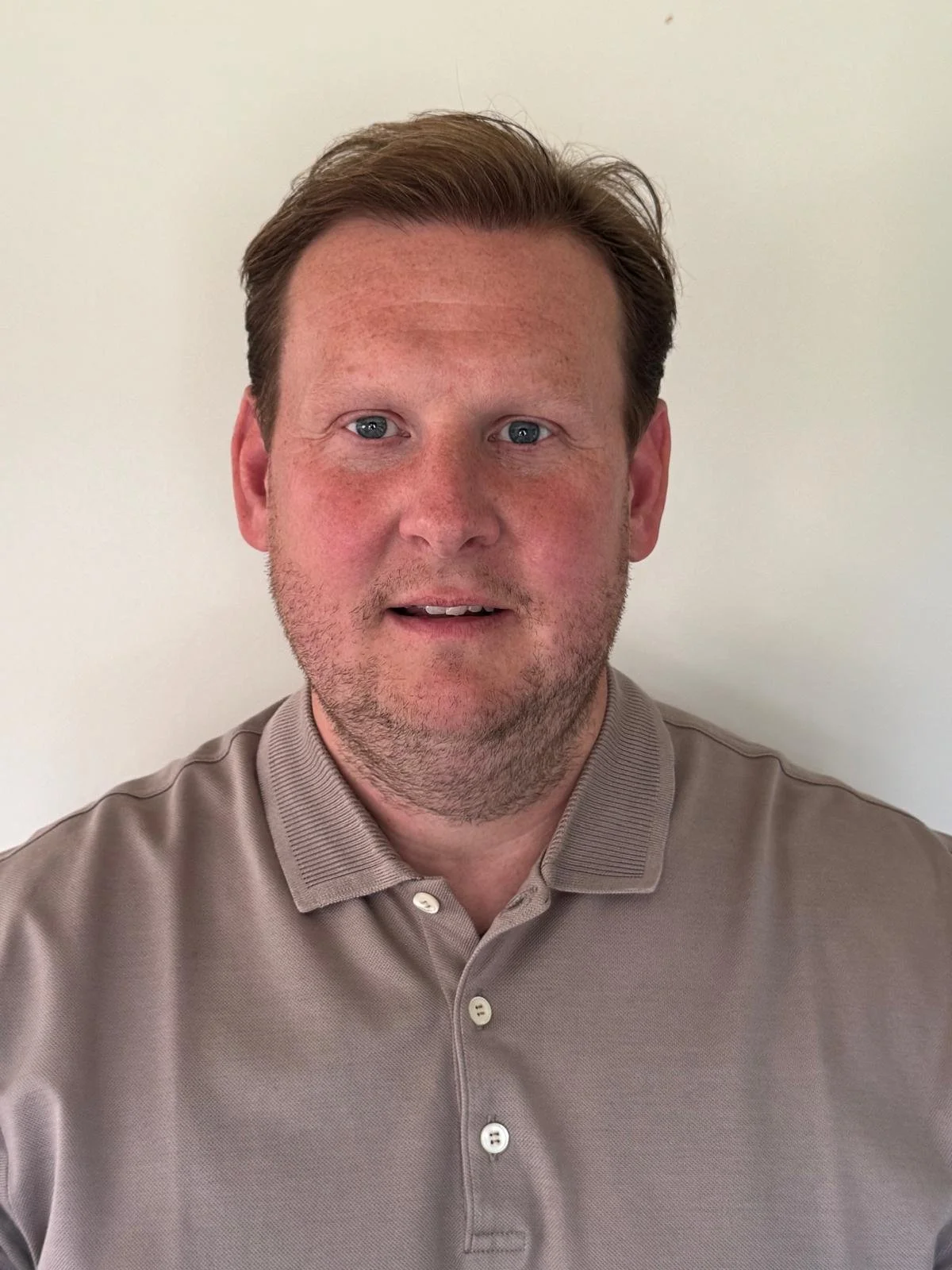 A man with reddish-brown hair and a light beard wearing a beige polo shirt, standing against a plain off-white background.