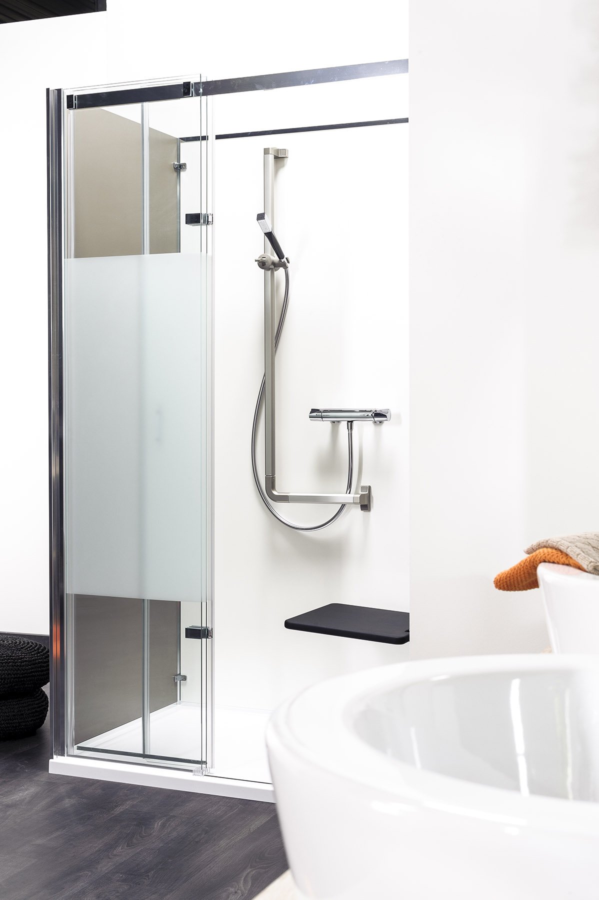Cabine de douche moderne avec une paroi en verre, une poignée de douche, une barre de fixation, une petite plateforme noire, dans une salle de bain blanche, avec des serviettes en orange et beige à droite.