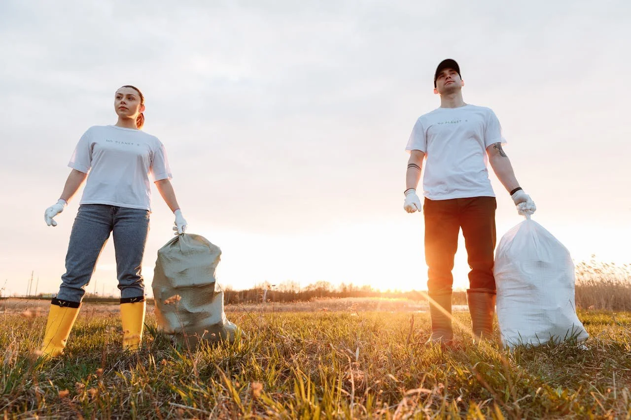 Two people holding trash bags and wearing gloves, cleaning up a field during sunset.