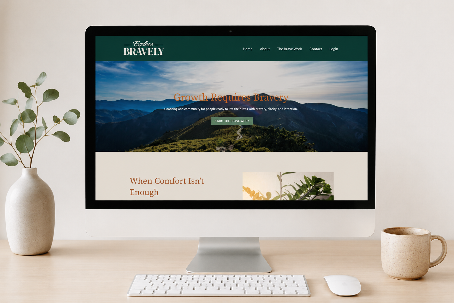 Computer monitor displaying a website titled 'Explore Bravely' with a scenic mountain landscape background, a plant in a vase to the left, a mug to the right, and a keyboard and mouse in front.