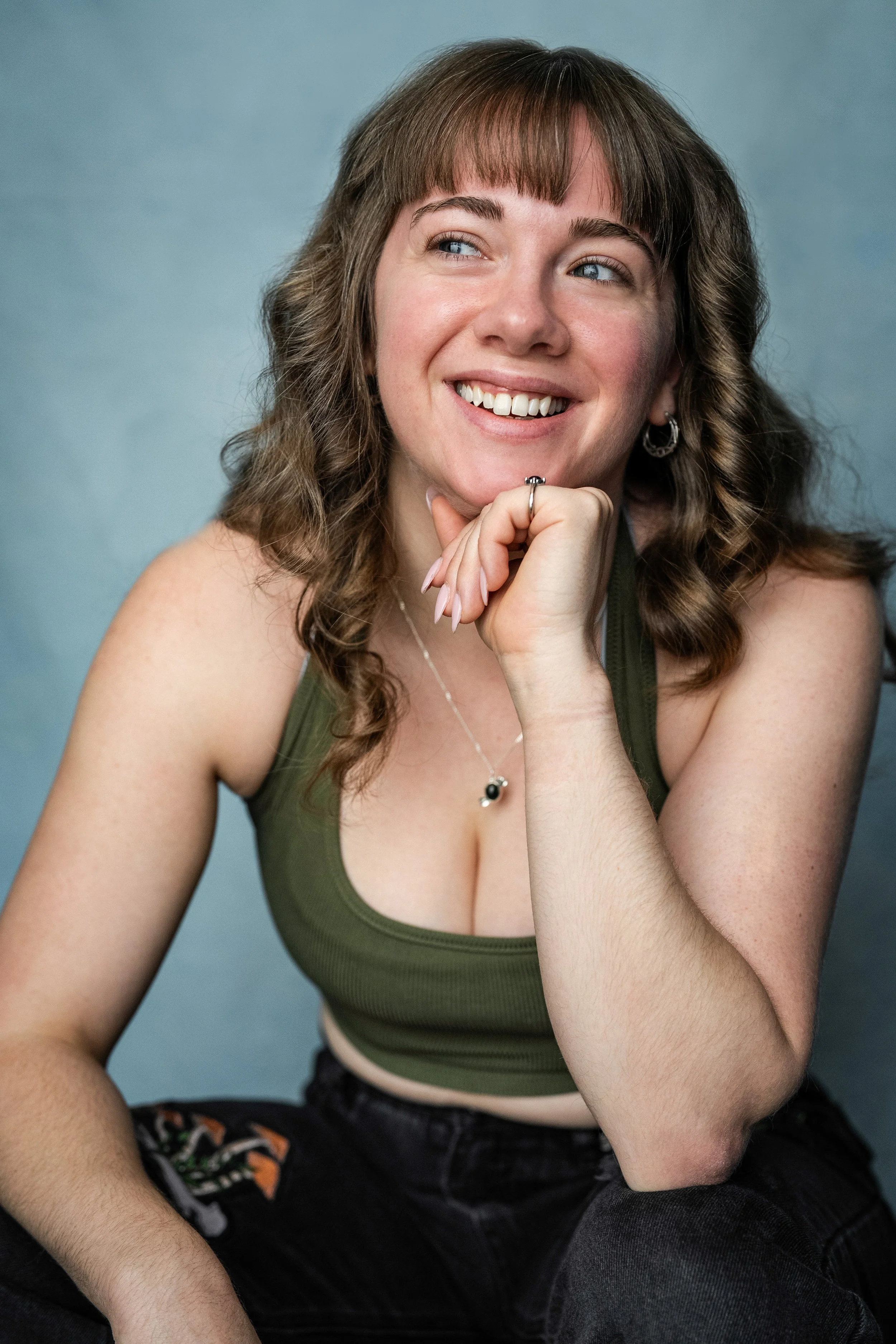 A smiling young woman with wavy brown hair, wearing a green sleeveless top and jewelry, sitting with her chin resting on her hand, looking to her left.