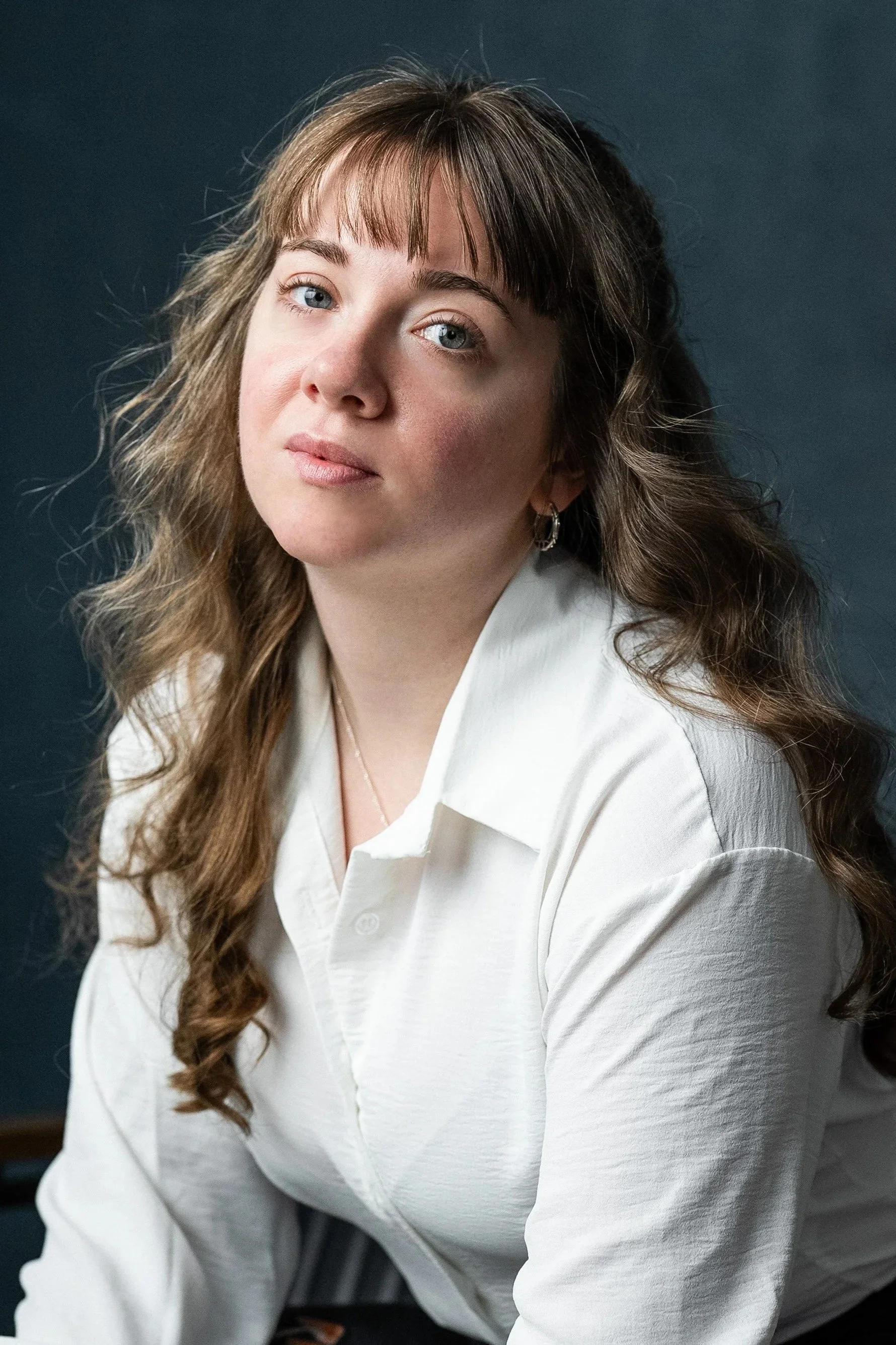 A woman with wavy brown hair, wearing a white shirt, posing for a portrait against a dark background.
