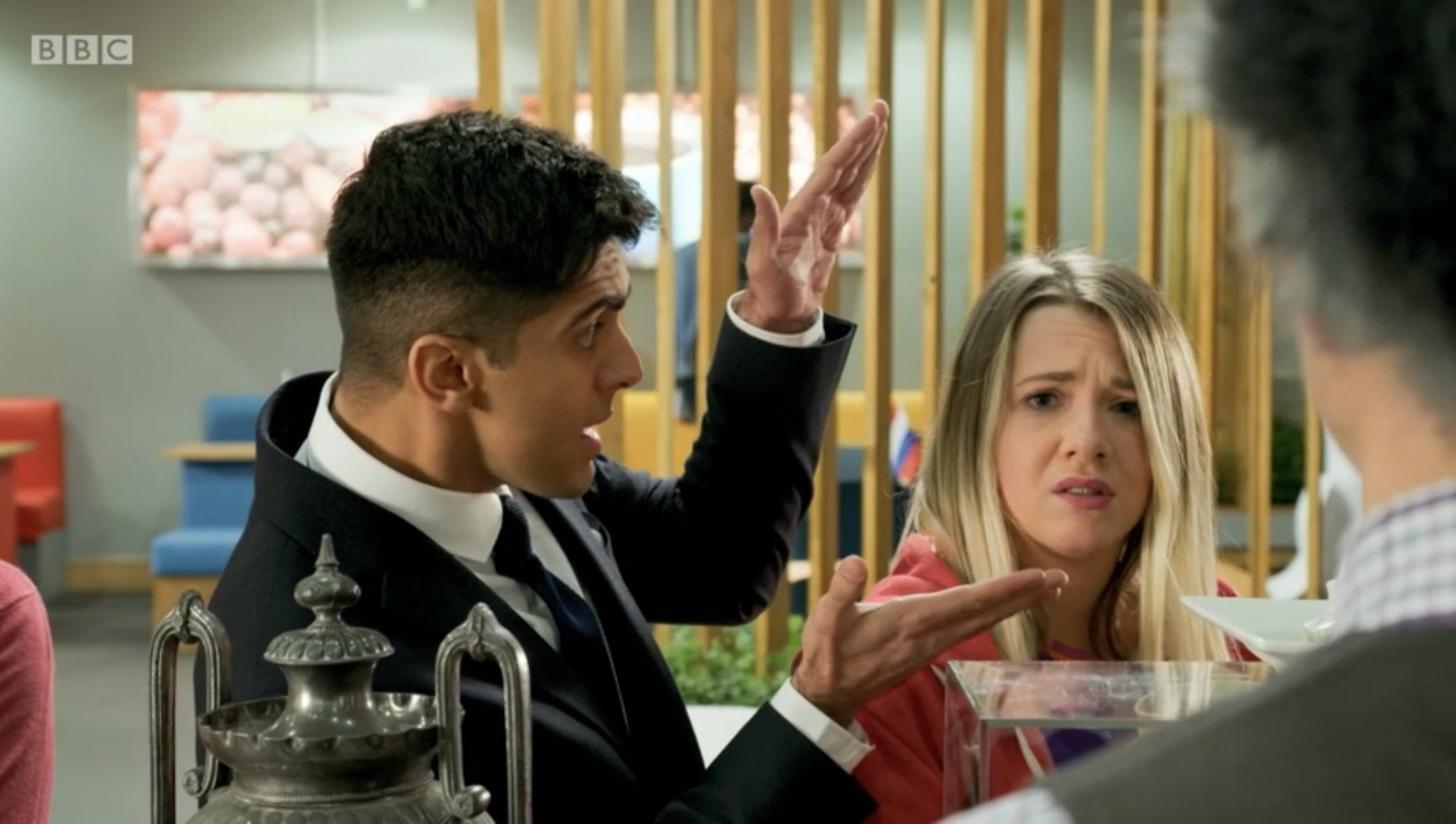A man in a black suit with a white shirt and black tie gestures with his hands while speaking, with a woman in a red jacket looking confused, and another person with dark hair seen from behind in the foreground.