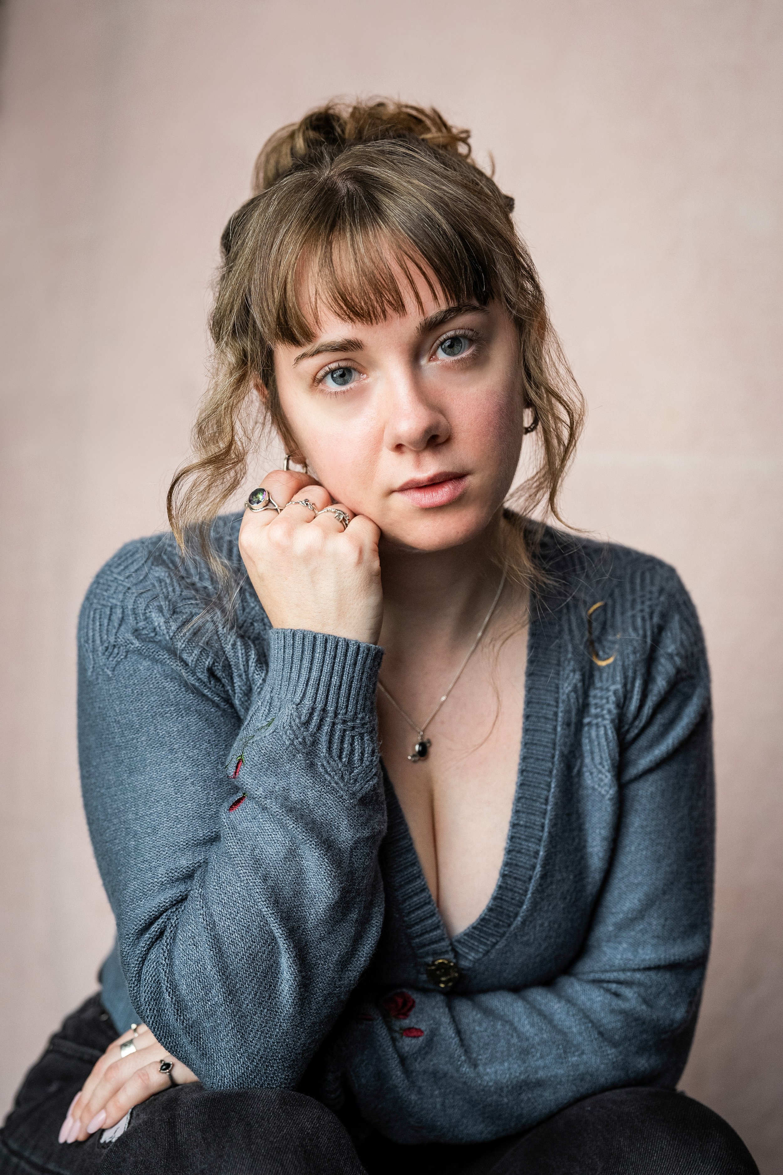 A young woman with blue eyes and light brown hair with bangs, wearing a gray cardigan, sitting against a beige background.