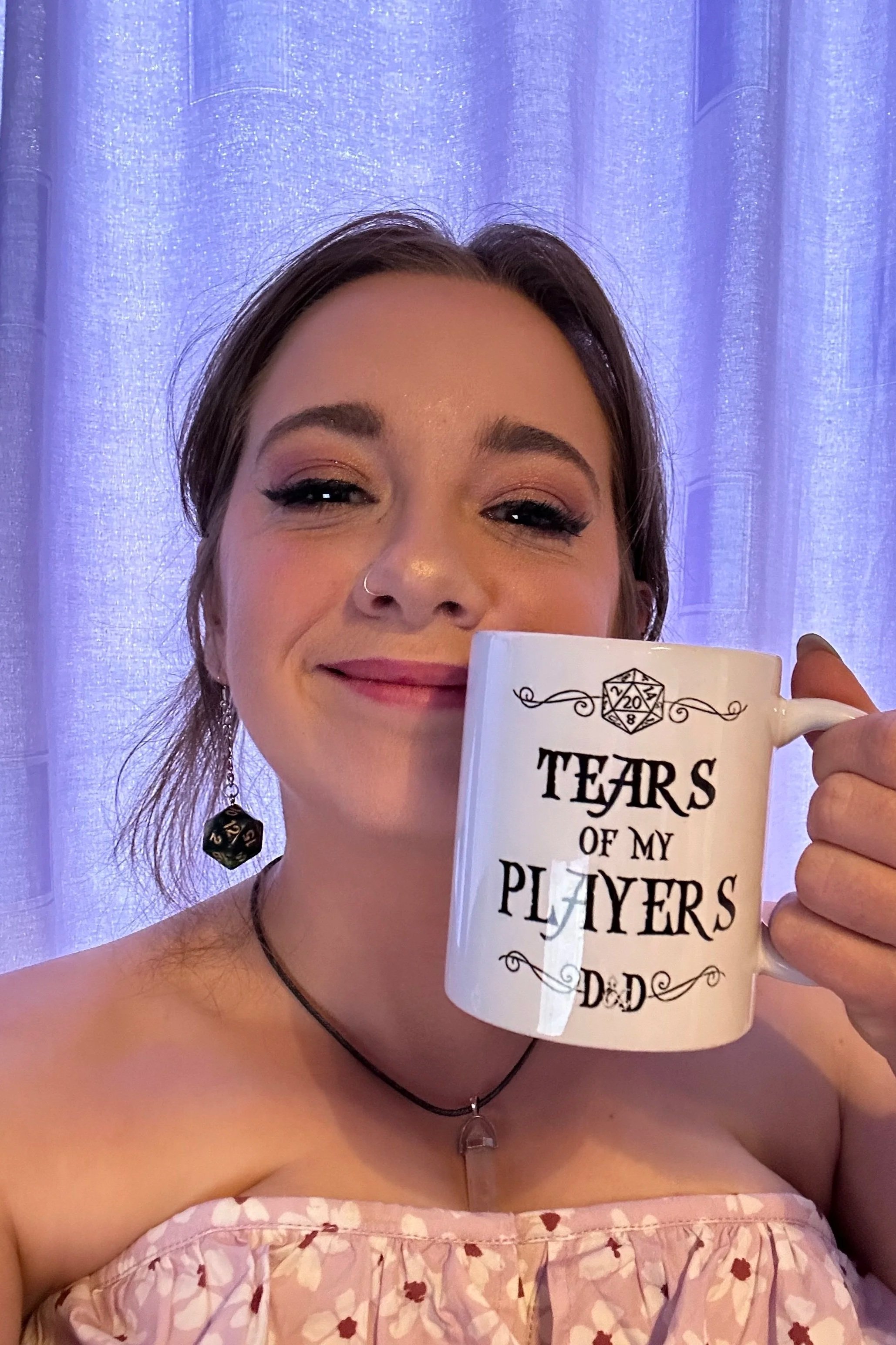 A woman holding a coffee mug that says 'Tears of my Players Dad' smiling at the camera, with purple curtains in the background.