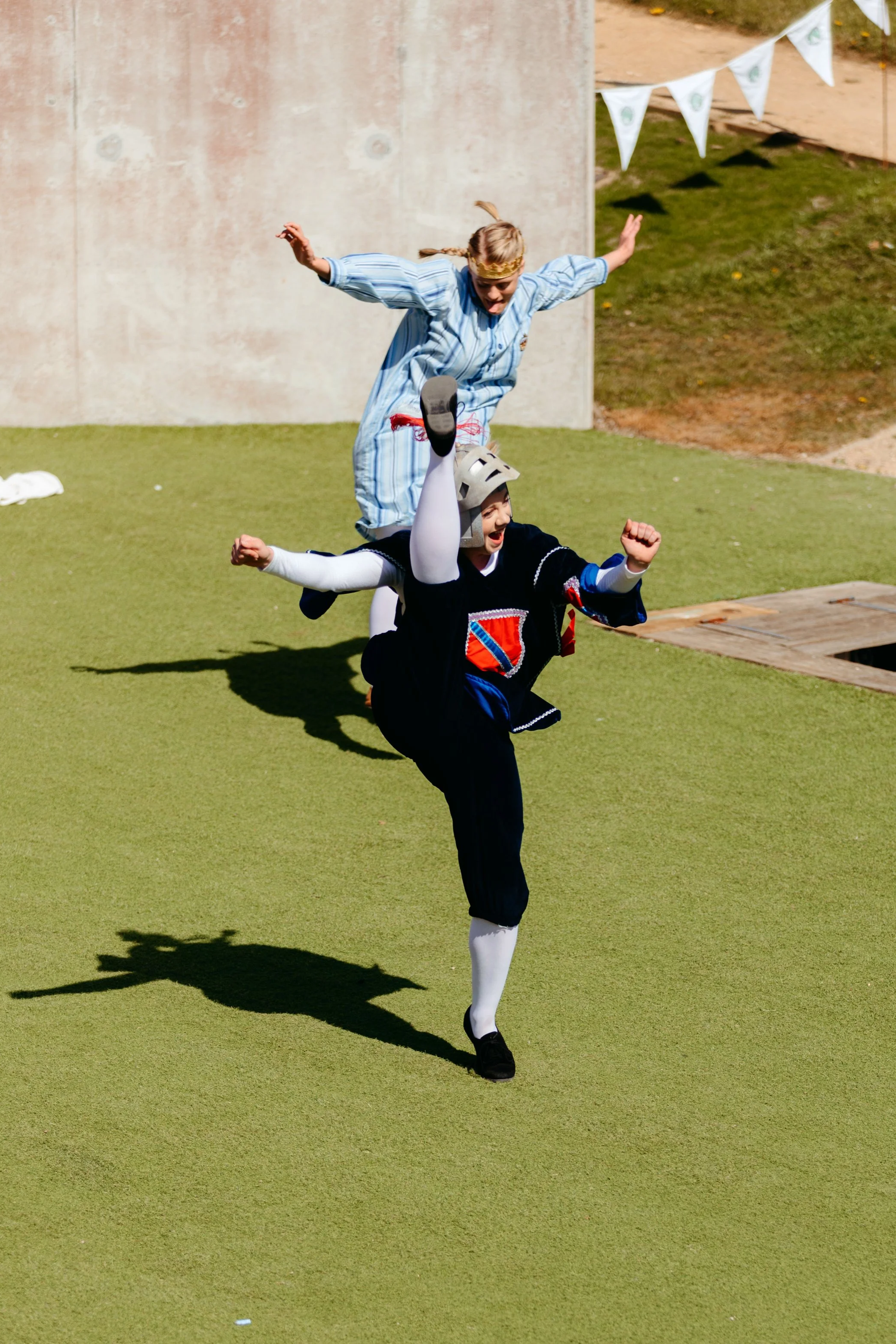 Two people dressed as theatrical characters are performing a playful acrobatic act outdoors on a grassy area. The person in the front is wearing a Knight costume with a helmet, a black tunic with a red and blue shield emblem, white tights, and black shoes. The person behind is dressed in a blue nightgown with a golden headband, mimicking a sleepy or surprised expression while balancing on the other’s back. Both are raising their arms and appear energetic and joyful.